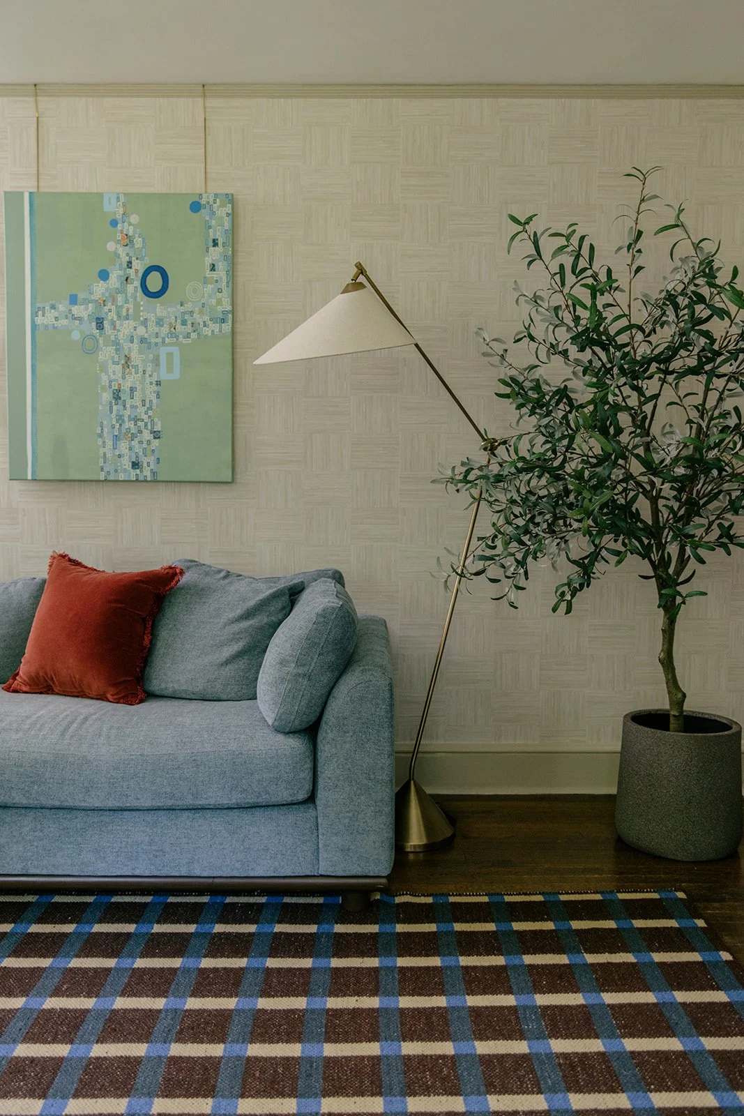 Living room with light blue sofa, rust-colored pillow, floor lamp, large potted tree, abstract artwork, patterned rug, and beige textured wallpaper.