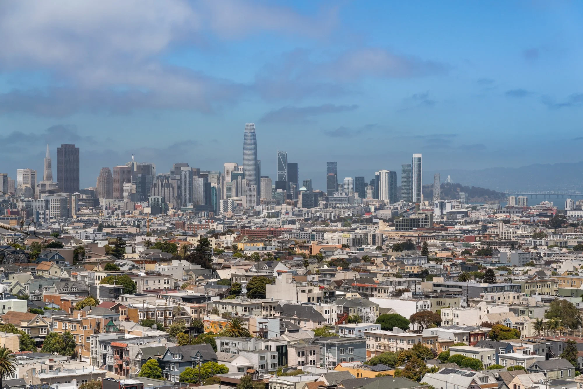 A panoramic cityscape of San Francisco with numerous residential houses in the foreground, modern high-rise buildings in the middle, and iconic skyscrapers including the Salesforce Tower in the downtown skyline. Clear partly cloudy sky above with the San Francisco Bay and distant hills in the background.