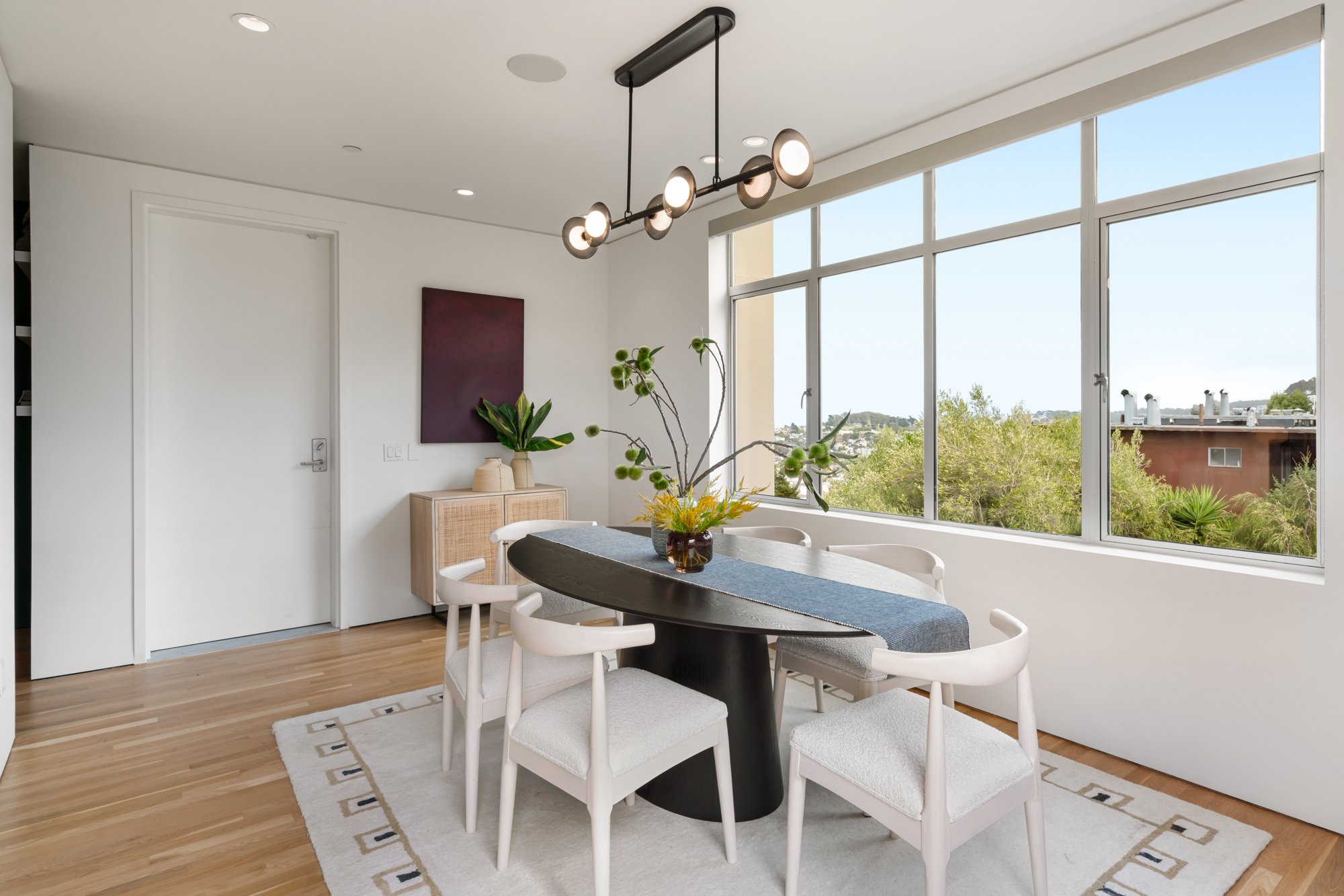 A modern dining area with a round black table, six white chairs, a gray table runner, and a glass vase with green and yellow flowers. Large windows show trees and a blue sky outside. A wooden sideboard with decorative vases and a large wall art piece