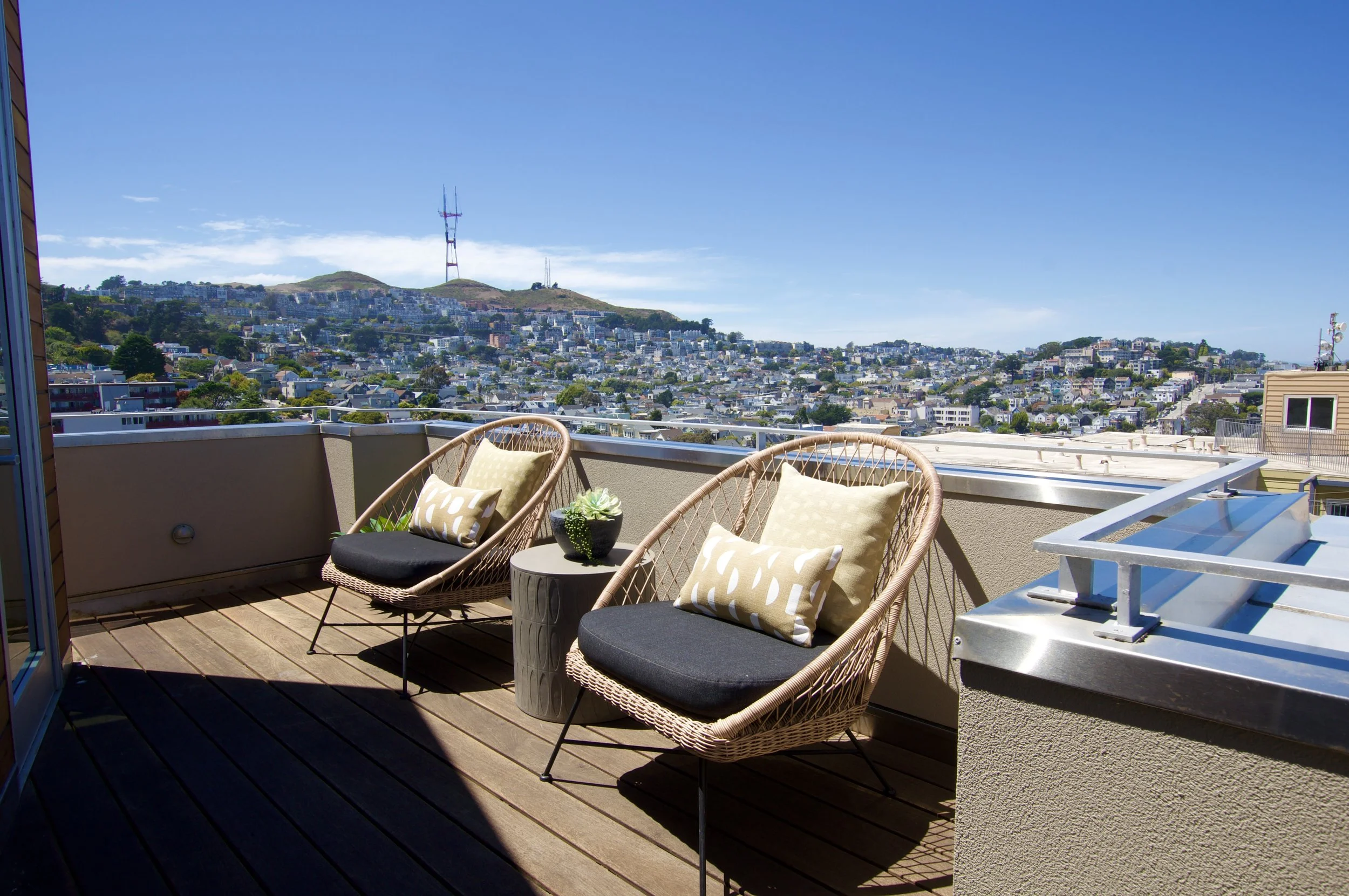 A rooftop balcony with two wicker chairs with black cushions and beige patterned pillows, a small round table with a plant, overlooking a cityscape with hilly terrain and a large radio tower in the background.