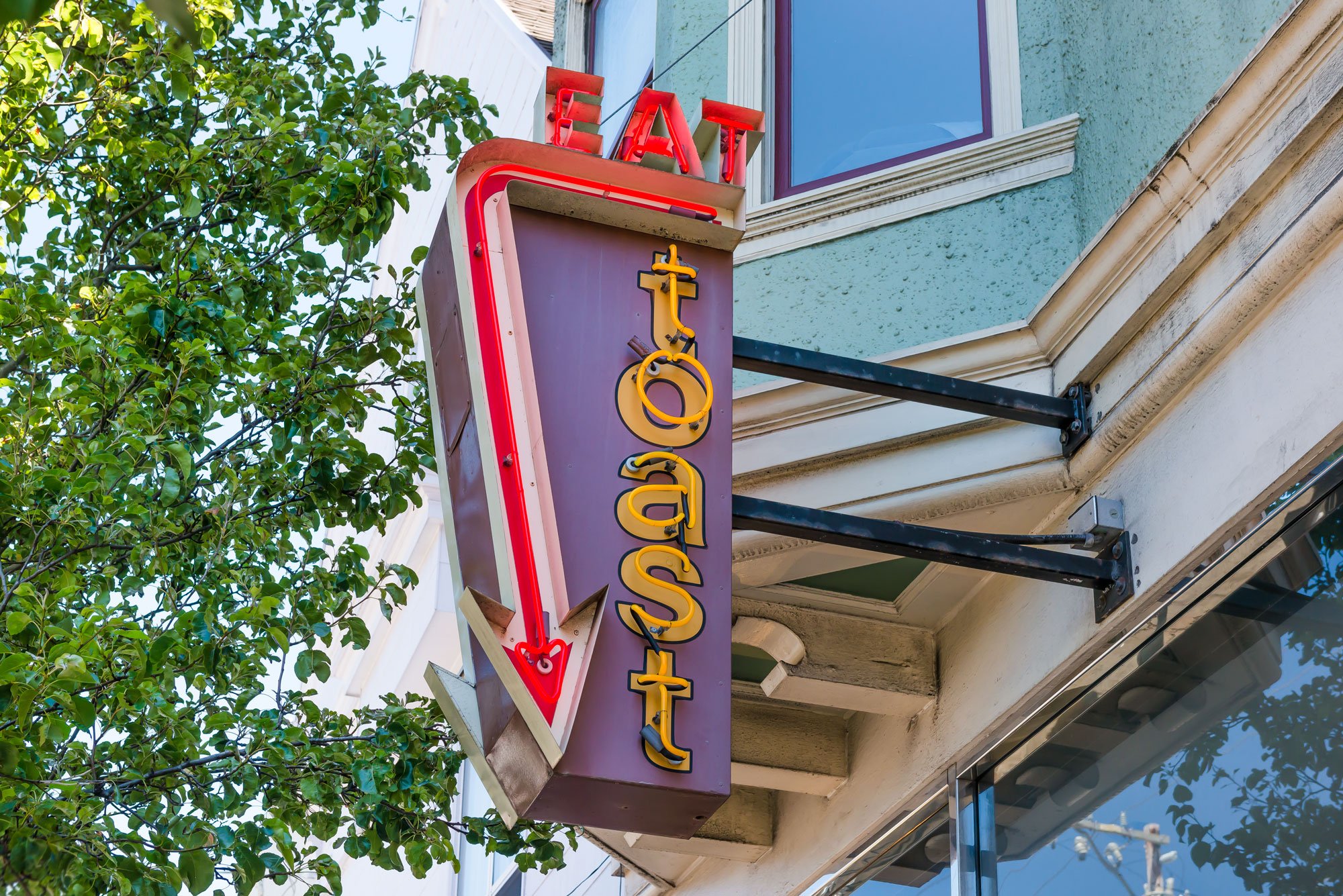 Neon sign for a tattoo shop with red upward arrow and yellow lettering, mounted on a building with a blue-green textured wall and surrounded by green trees.