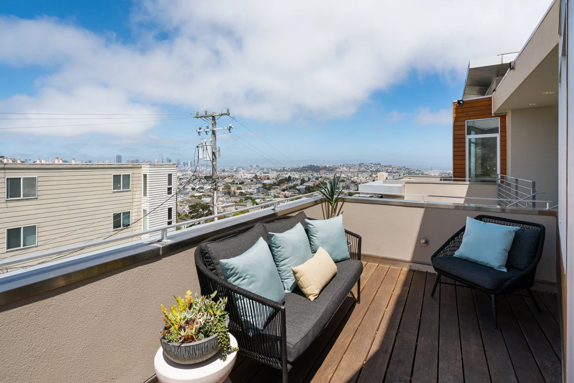 A balcony with outdoor seating including a black cushioned loveseat and a matching chair, each with pillows, and a white circular table with a potted succulent plant. The balcony overlooks a cityscape with numerous buildings, power lines, and a blue 