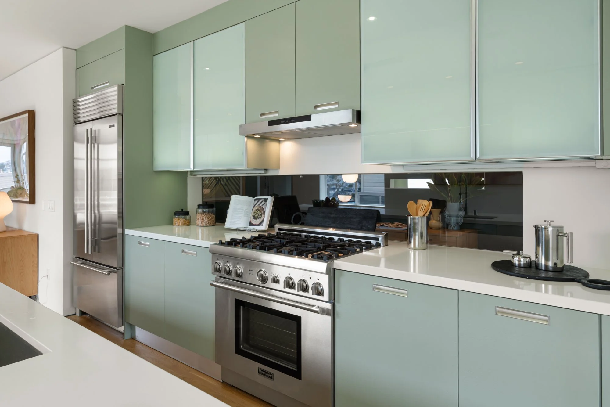 Modern kitchen with mint green cabinets, stainless steel appliances including a refrigerator and a gas oven, white countertops, and decorative jars on the counter.