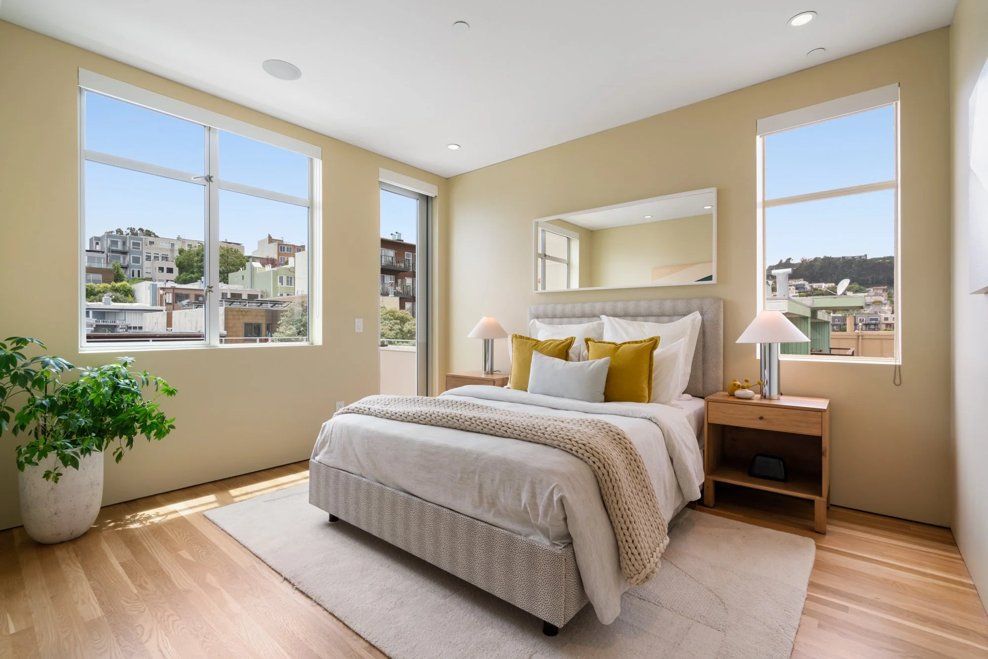 A bedroom with large windows showing city buildings and trees outside. The bed has white bedding with mustard-yellow and white pillows, and a beige blanket. There are two small wooden nightstands with lamps, a large mirror above the bed, and a potted