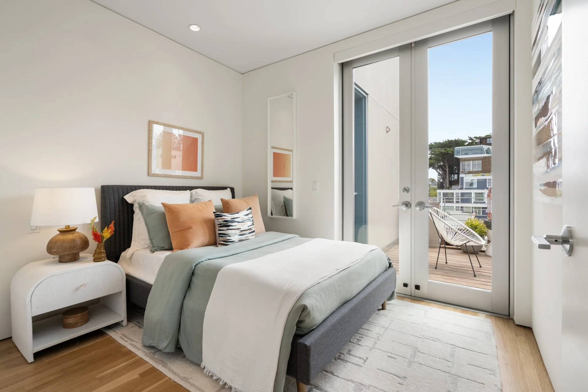 A bedroom with a bed made with white and pastel green bedding and throw pillows, a nightstand with a lamp and decorative items, and a large sliding glass door leading to a balcony with outdoor furniture, overlooking neighboring buildings and trees.