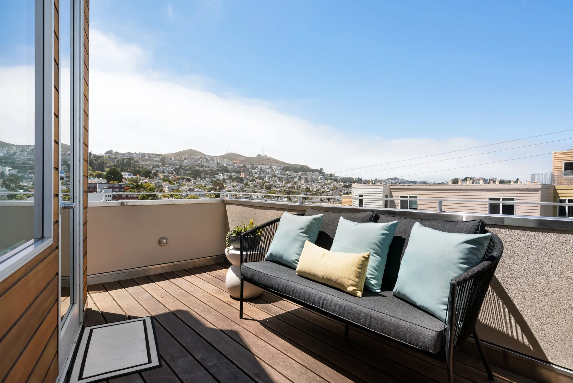 Balcony with outdoor seating area, including a dark gray cushioned sofa with pillows, potted plant, and view of hillside cityscape with houses and hills under a partly cloudy sky.