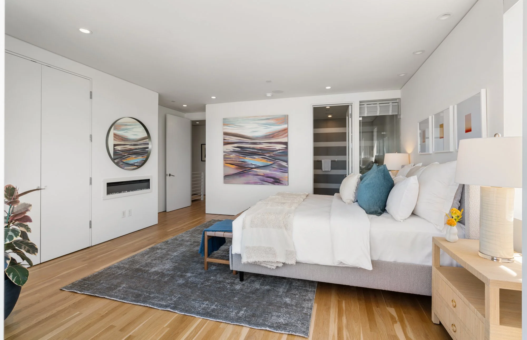 Modern bedroom with white walls, wooden floor, and neatly made bed with white bedding and blue pillows. Artwork on the wall, a bedside table with a lamp and flowers, closet doors, and a glass-walled bathroom are visible.