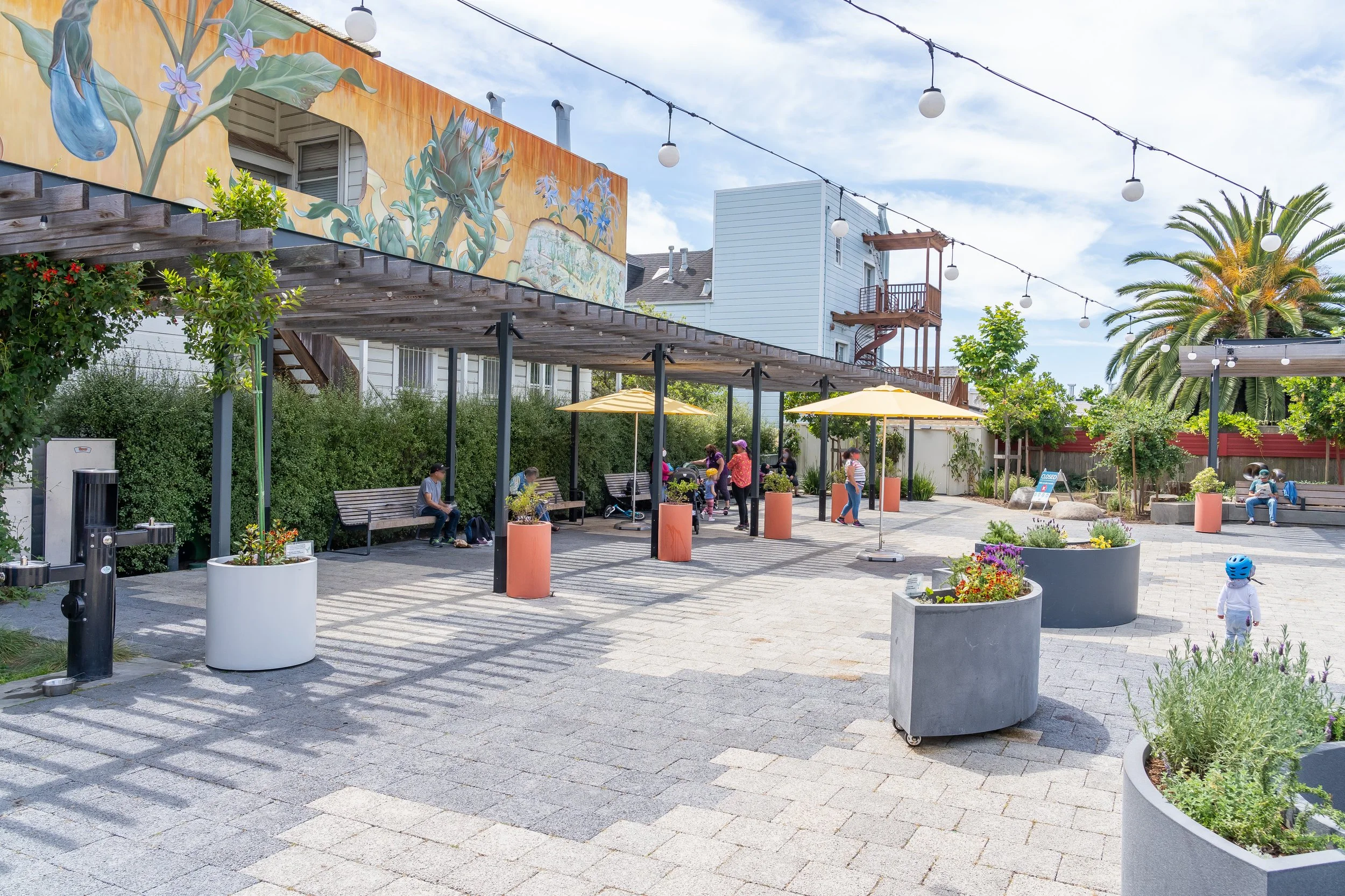An outdoor urban space with benches, large planters, trees, and string lights. A mural of plants, flowers, and a blue creature decorates the building in the background. People are sitting, chatting, and children are playing.