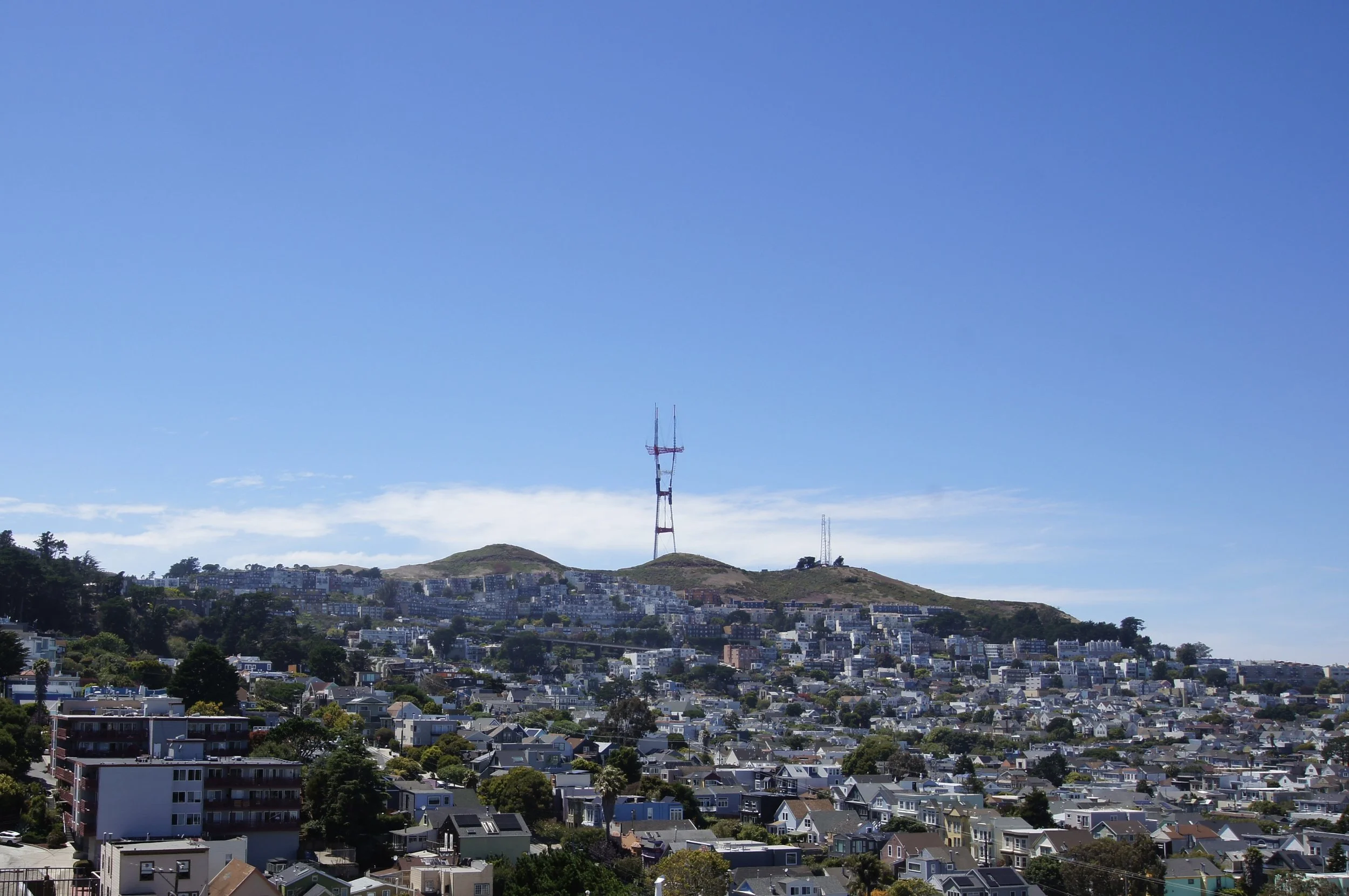 A cityscape with numerous residential houses and apartment buildings expanding towards hills in the background. The hills have several transmission towers, including a prominent tall tower with antennas in the center. The sky above is clear and brigh