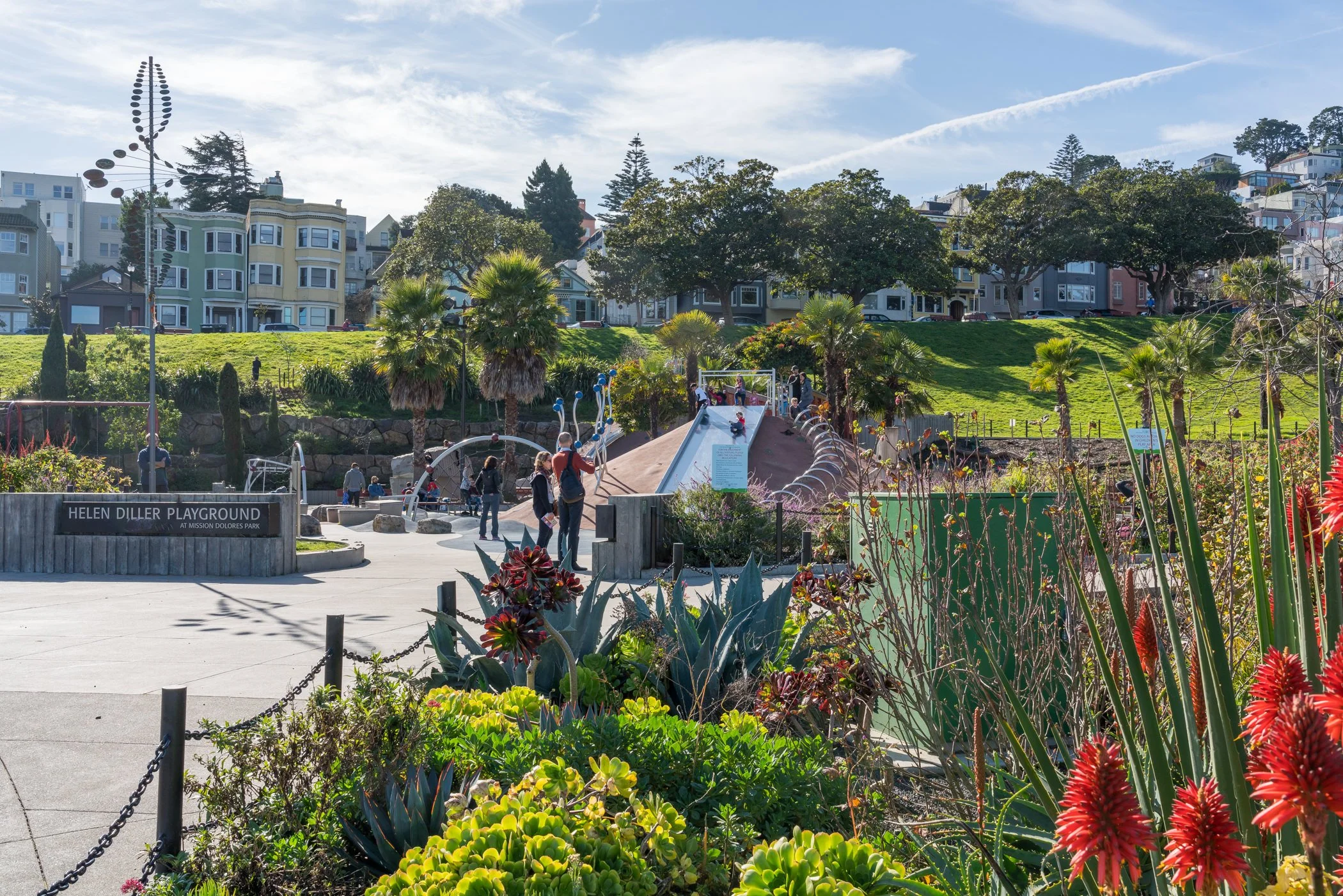 A playground with a slide, plants, and children, with colorful houses and trees on a hill in the background under a blue sky with light clouds.