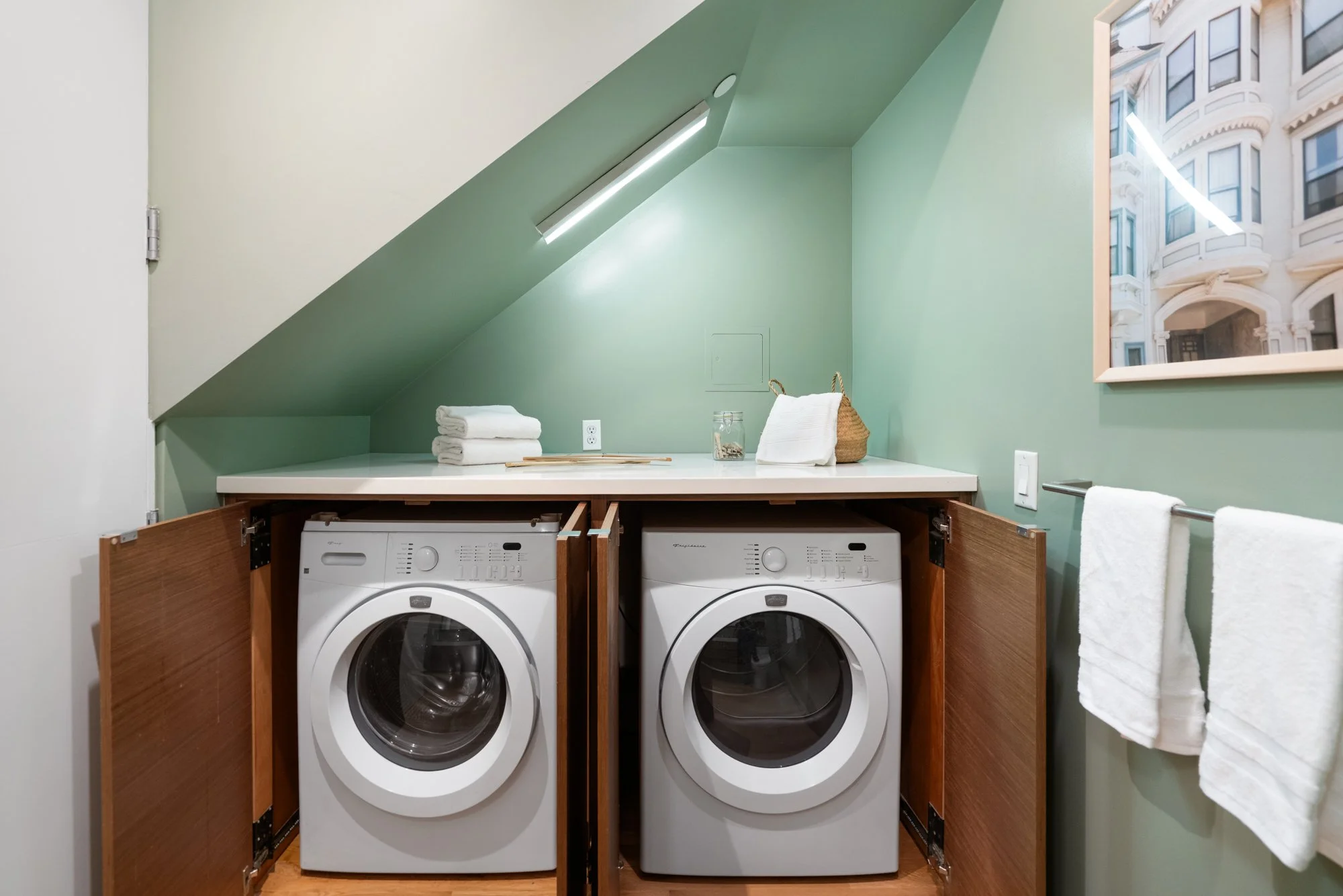 A laundry room with a green angled ceiling, a window showing a cityscape, and washing machines in a wooden cabinet. There are towels, a glass jar, and a basket on the counter.