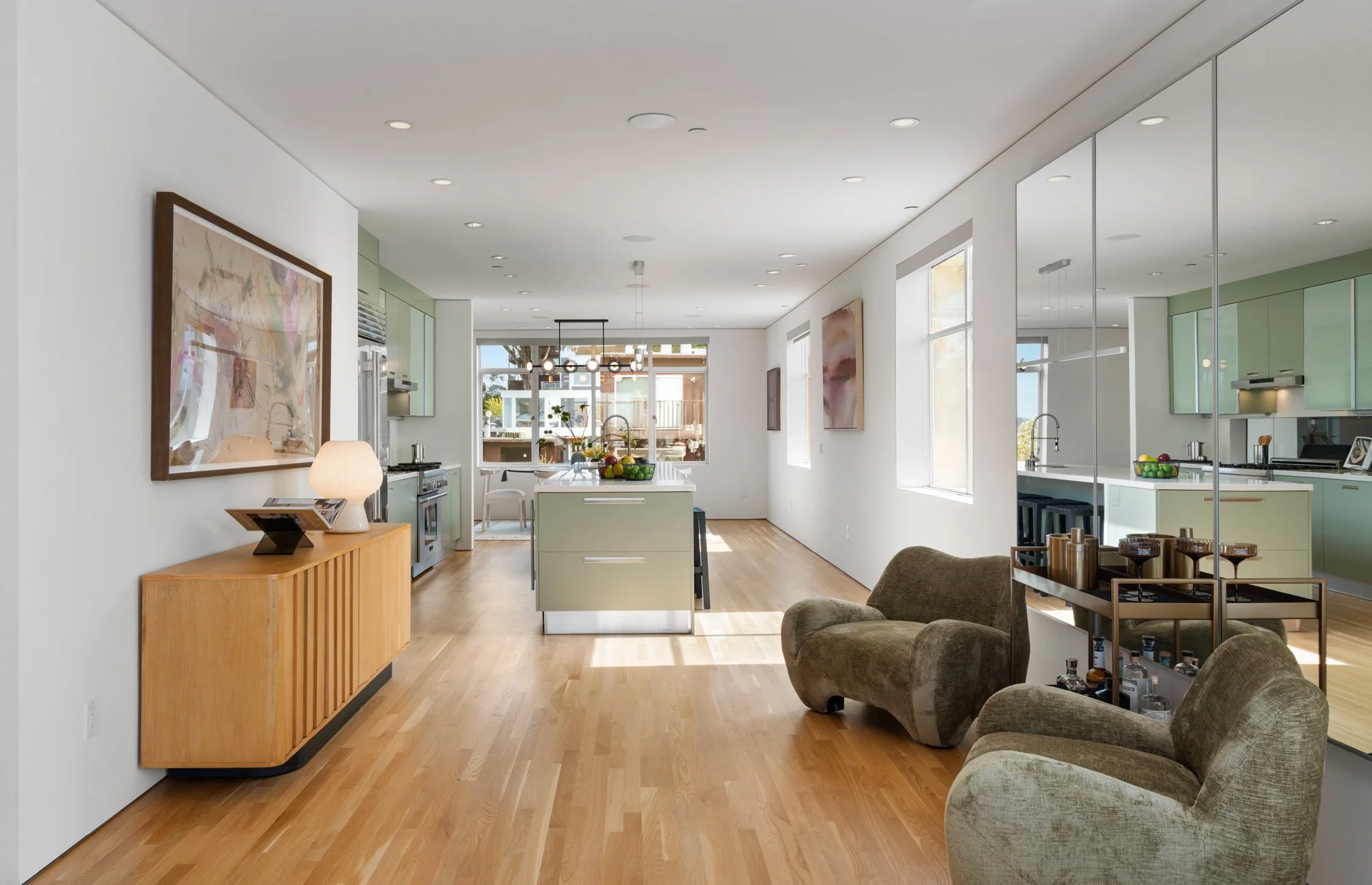 Modern open-concept kitchen and living room with minimalistic decor, hardwood floors, large windows, and art on the walls.