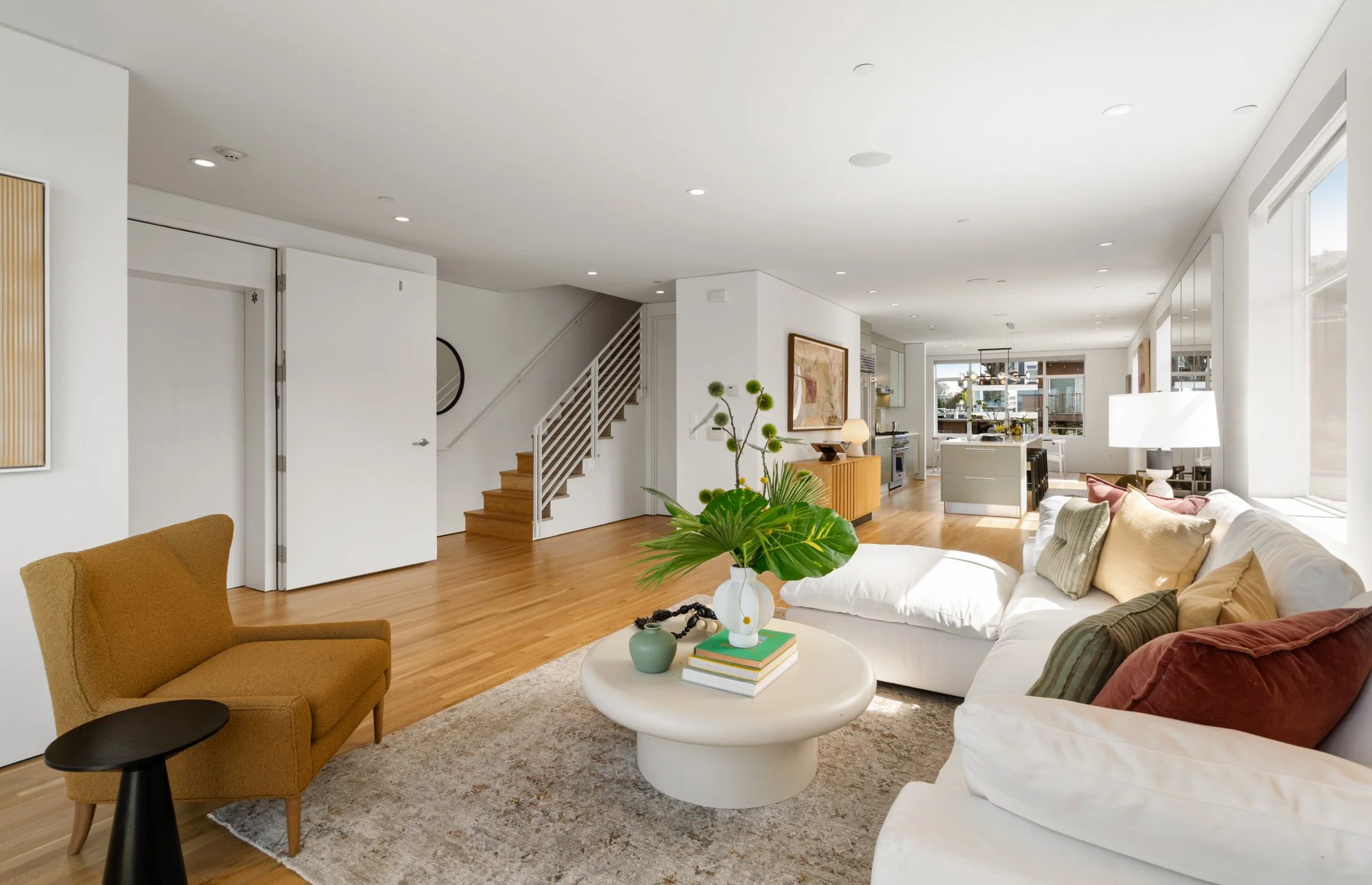 Spacious living room with white walls and ceiling, hardwood floors, a large white sofa with colorful pillows, a mustard armchair, a round coffee table with plants and books, and a view of the kitchen and dining area in the background. Large windows l