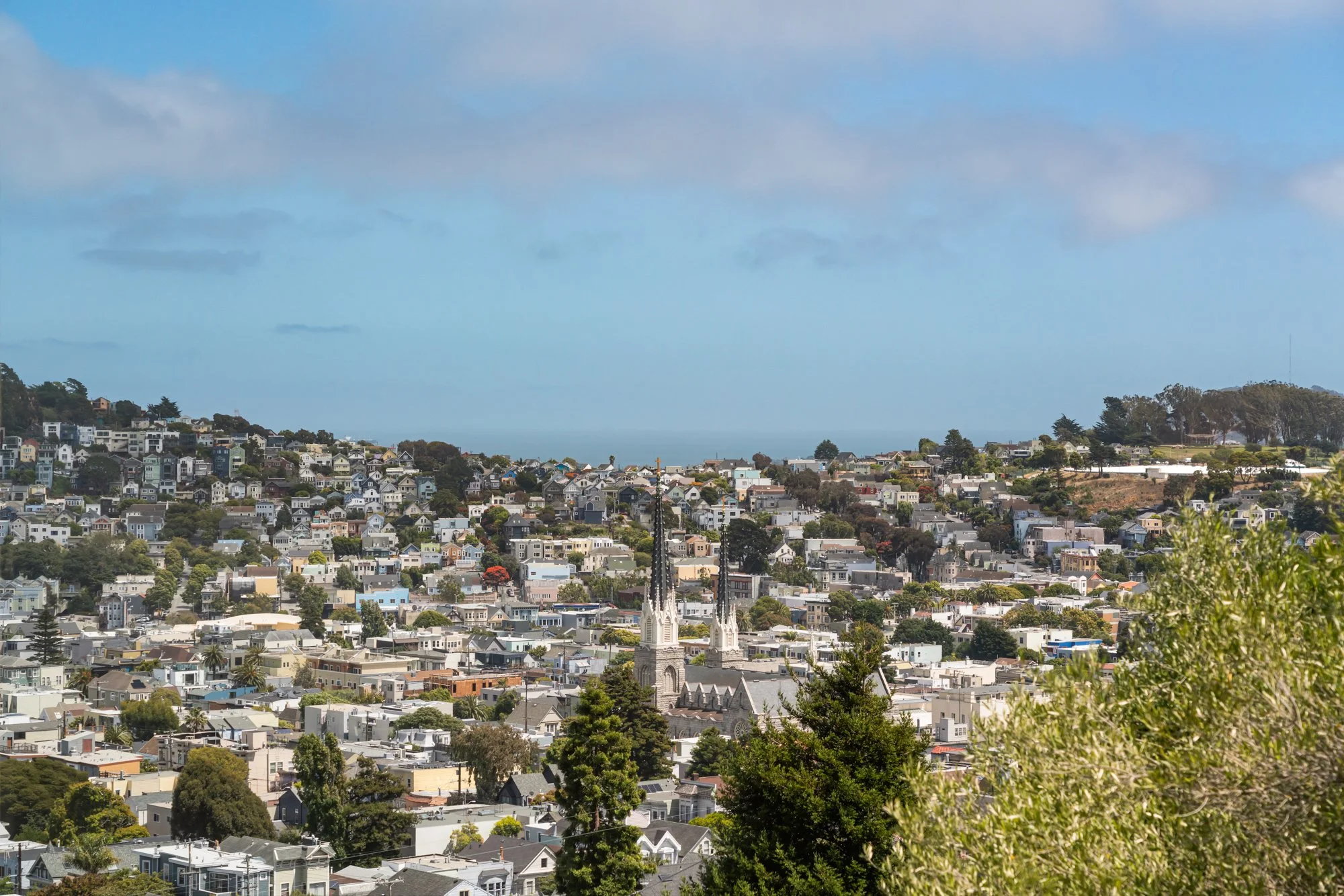 A cityscape of a residential neighborhood with numerous houses, trees, and a church with twin steeples in the foreground, hillside homes in the background, and blue sky with scattered clouds overhead.