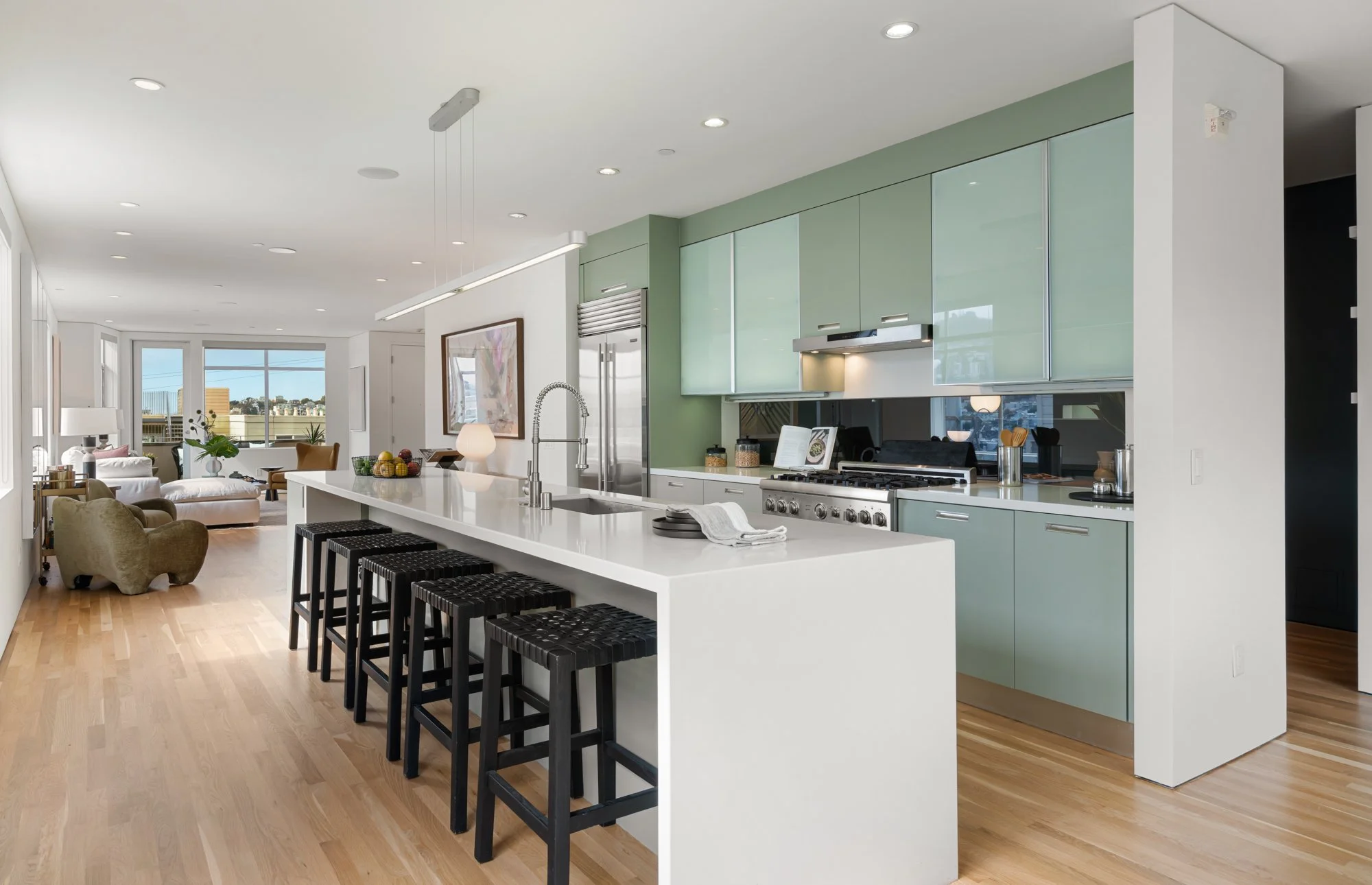 Modern open-concept kitchen with light green cabinets, white island, black stools, stainless steel appliances, and a spacious living area with large windows.