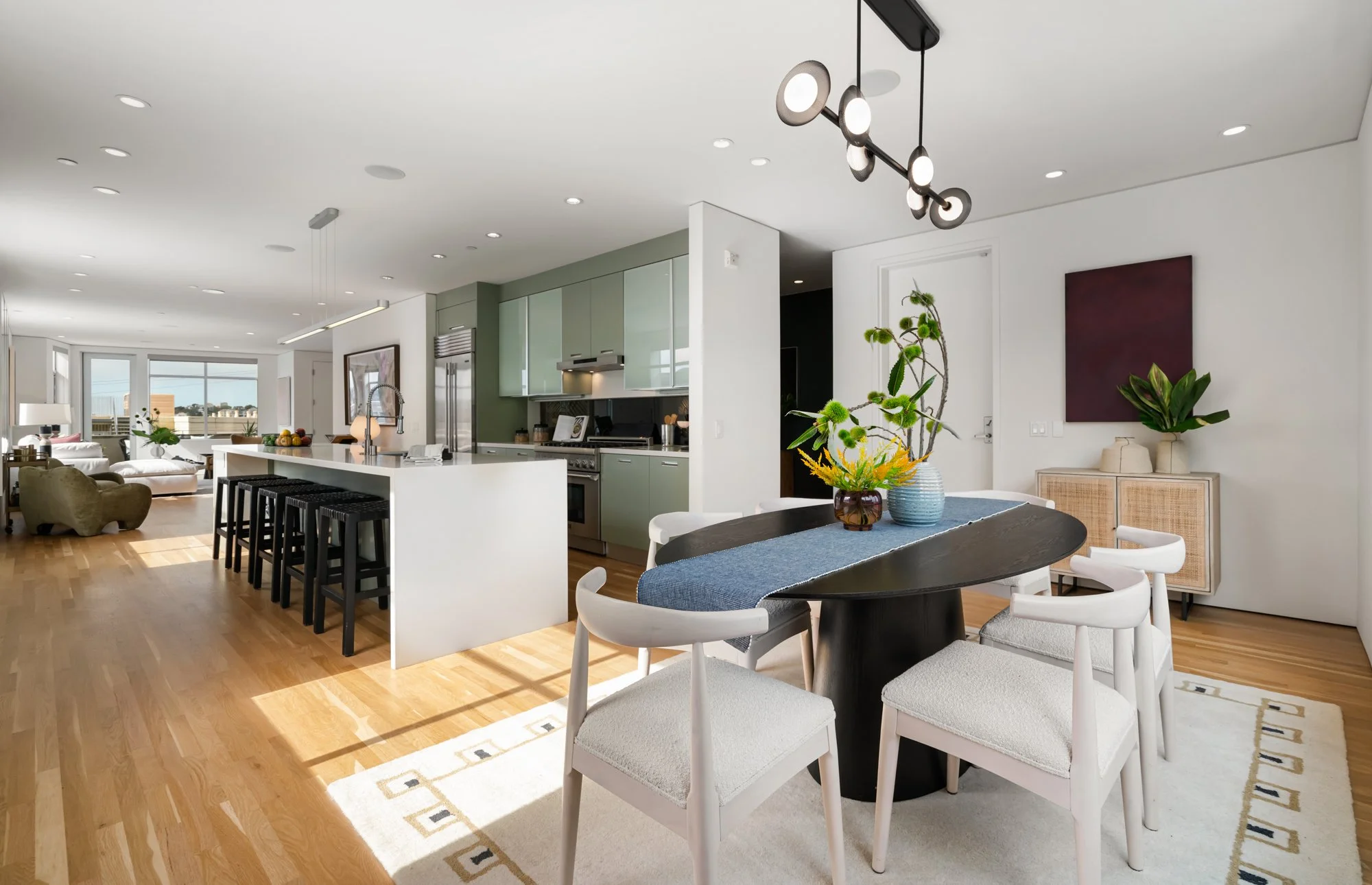 Open-concept living and dining area with a kitchen in the background. There is a wooden floor, a white rug, white dining chairs around a black table, and a decorative vase with green plants on the table. The kitchen has a white island with black stoo