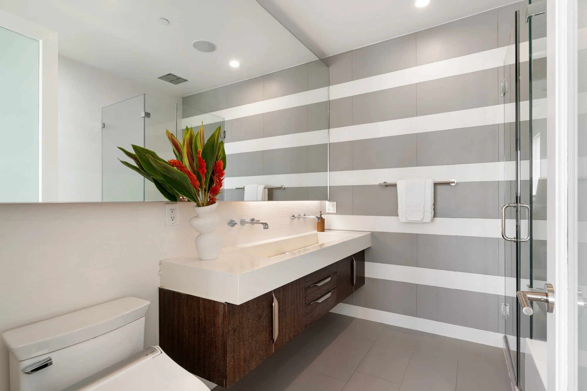 Modern bathroom with gray and white striped wall, glass shower enclosure, white countertop with faucet, wooden vanity, decorative plant in white vase, and towels.