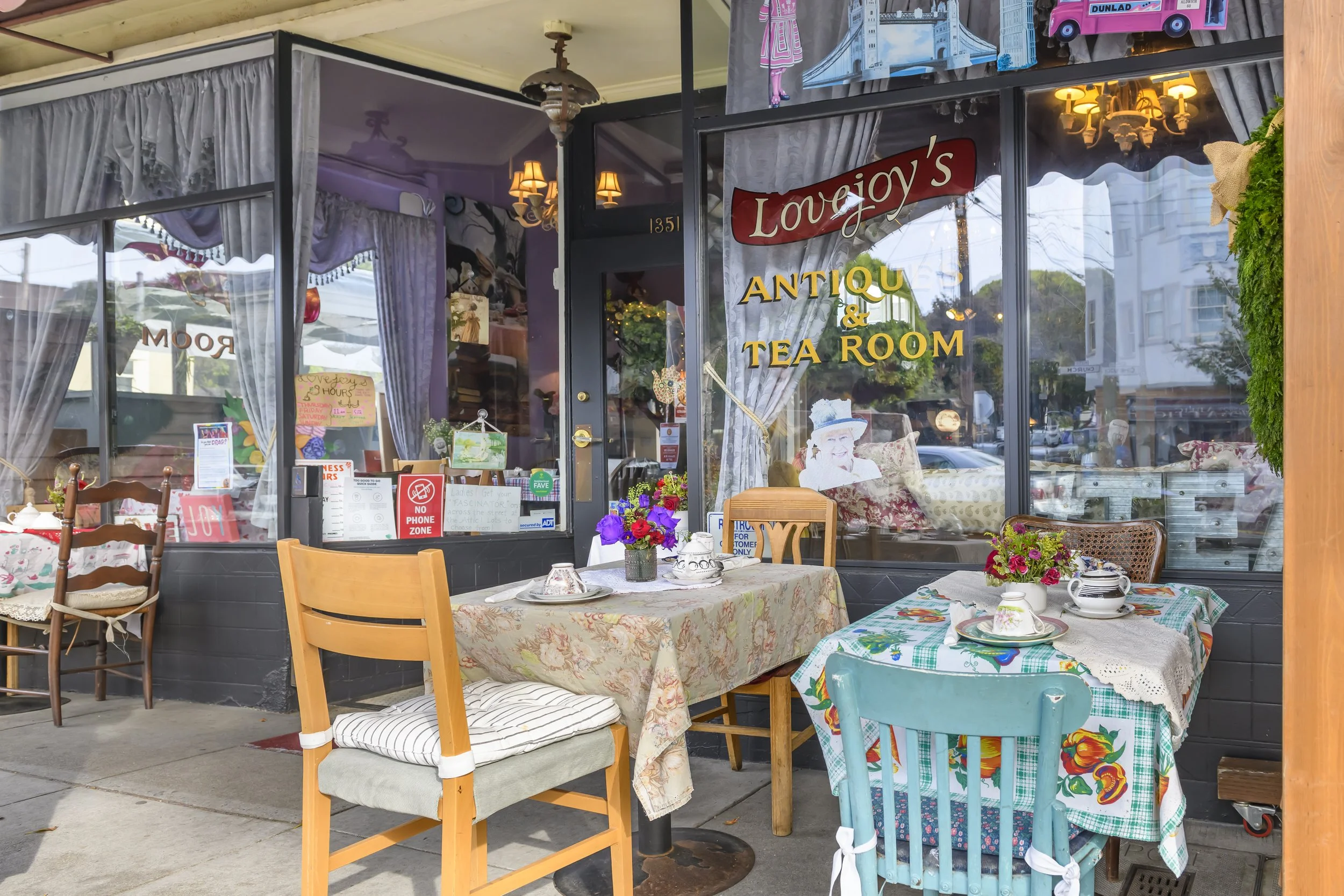 The storefront of Lovejoy's Antiques & Tea Room with tables set for dining outside, decorated with colorful tablecloths, flowers, and small decorative items.