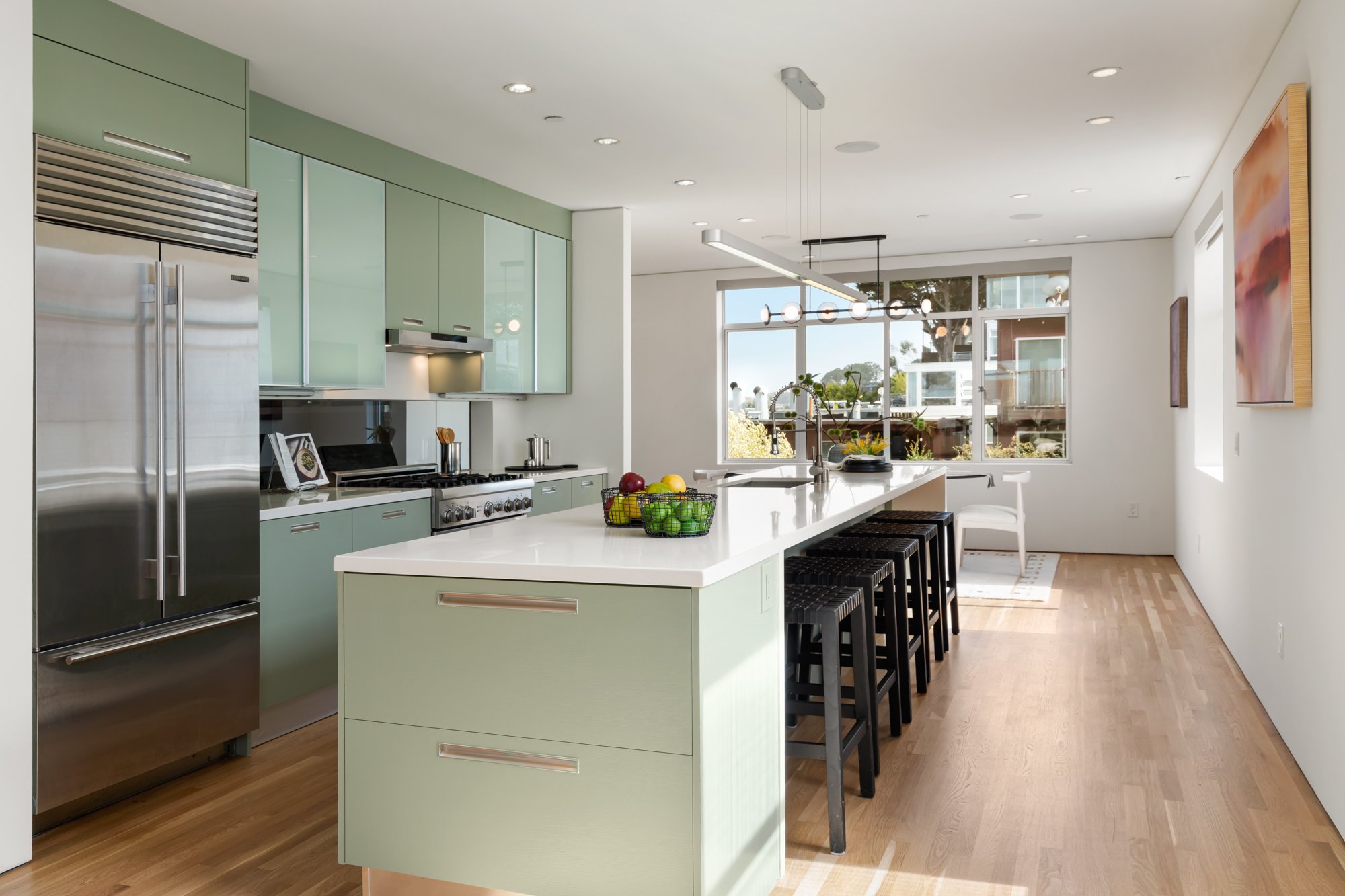 Modern kitchen with pale green cabinetry, stainless steel appliances, a large white island with black barstools, and large windows showing outdoor trees.