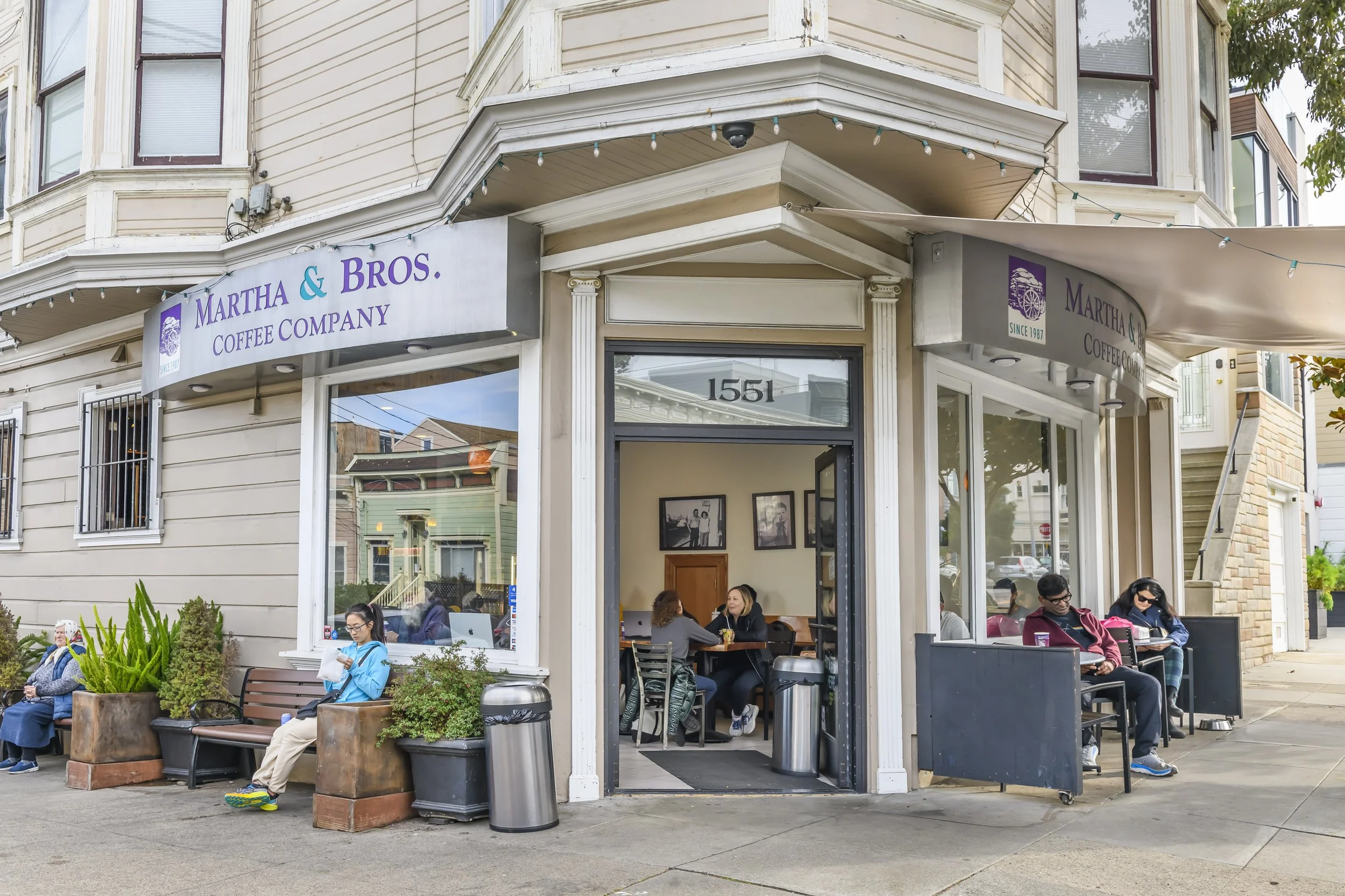 Exterior of Martha & Bros. Coffee Company with customers sitting outside, some using laptops, on a city sidewalk during daytime.