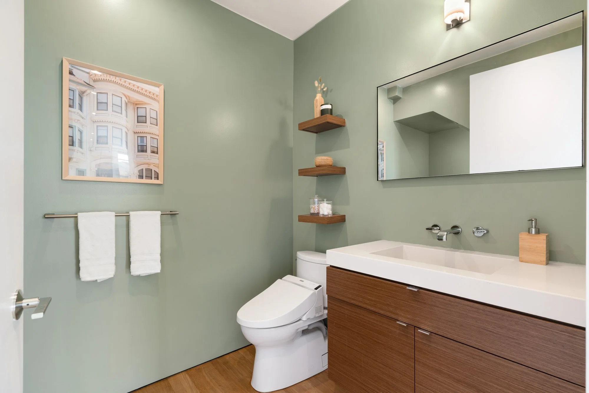 Modern bathroom with light green walls, a large mirror, wooden shelves with decorative items, towel rack with two white towels, a toilet, and a sink vanity with drawers, a soap dispenser, and a brown tiled floor.