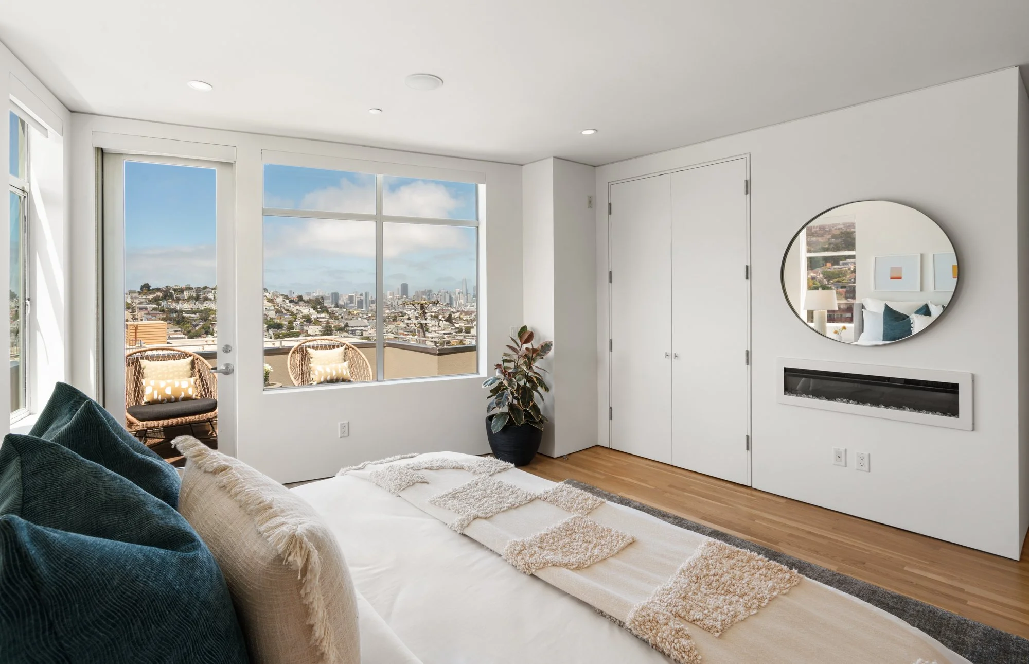 Modern living room with large window view of cityscape, beige sofa with pillows, potted plant, round mirror, and fireplace.