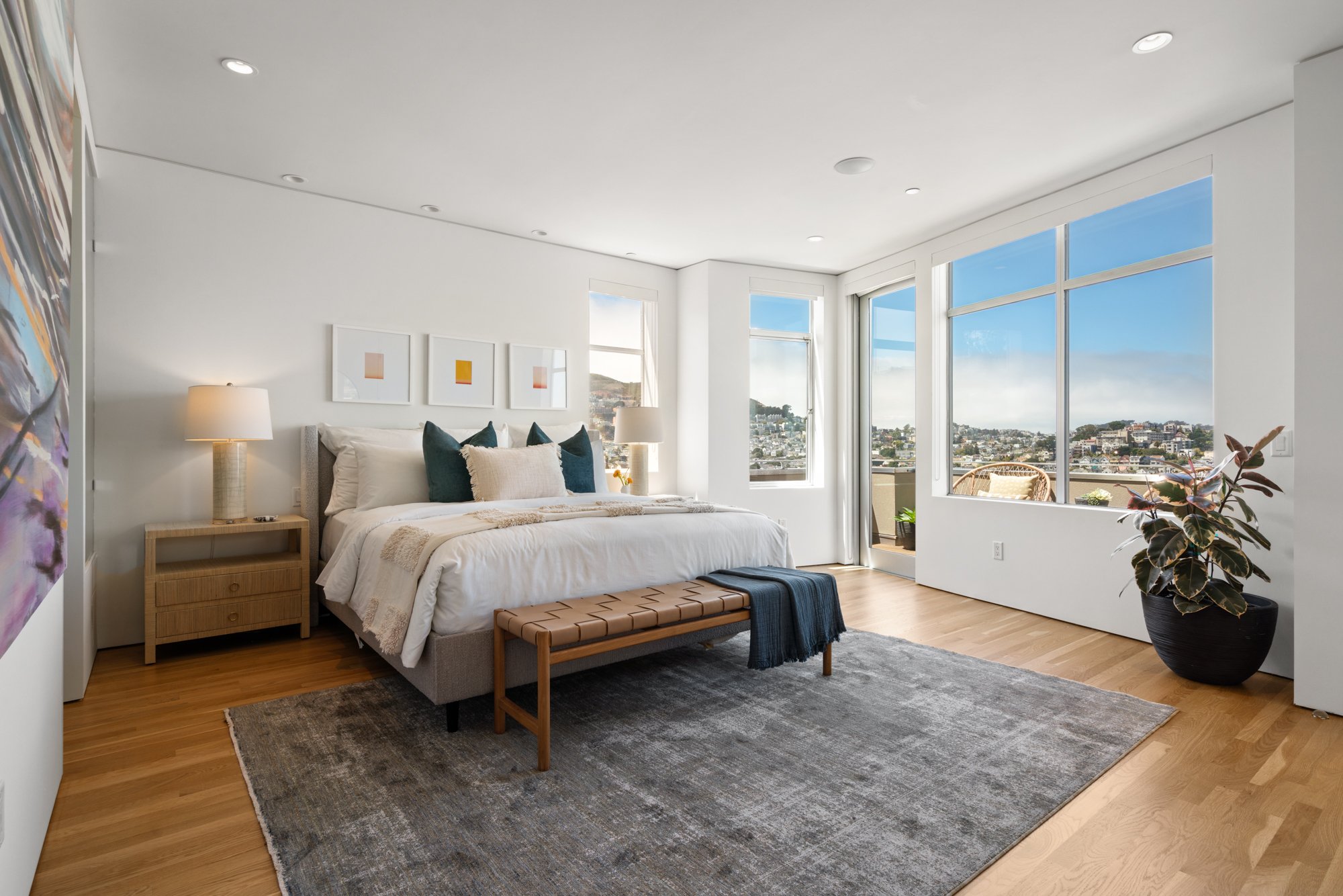 Bright bedroom with large windows showing city view, with a bed, nightstands, lamps, colorful pillows, artwork, and a potted plant.