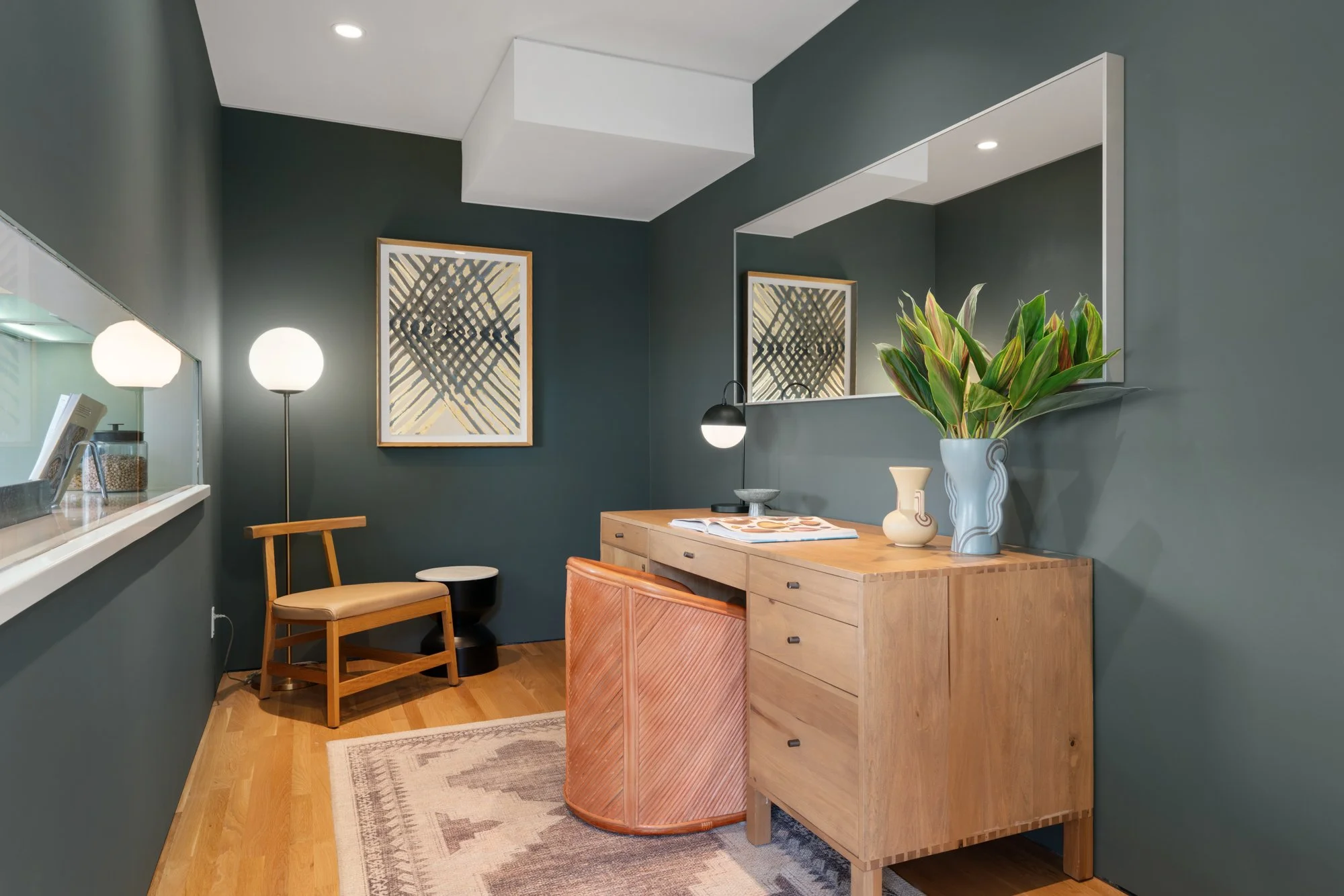 Modern dining room with dark teal walls, wooden sideboard, large mirror, and green leafy plant. Artistic framed art, white ceiling, and warm wooden floor.