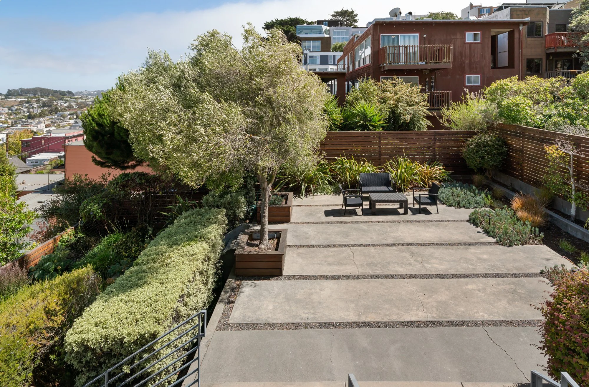 View of a terrace garden with outdoor seating, surrounded by lush greenery and plants, with residential buildings on a hillside in the background under a partly cloudy sky.