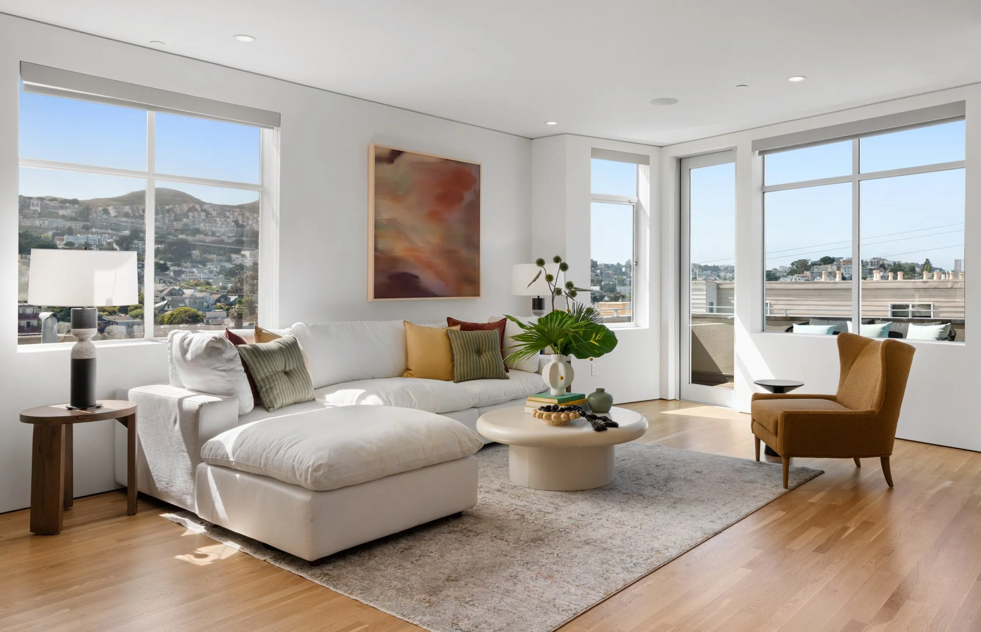 Bright living room with large windows, white sofa with pillows, a brown armchair, round coffee table with decorative items, and abstract wall art, with a city view outside.