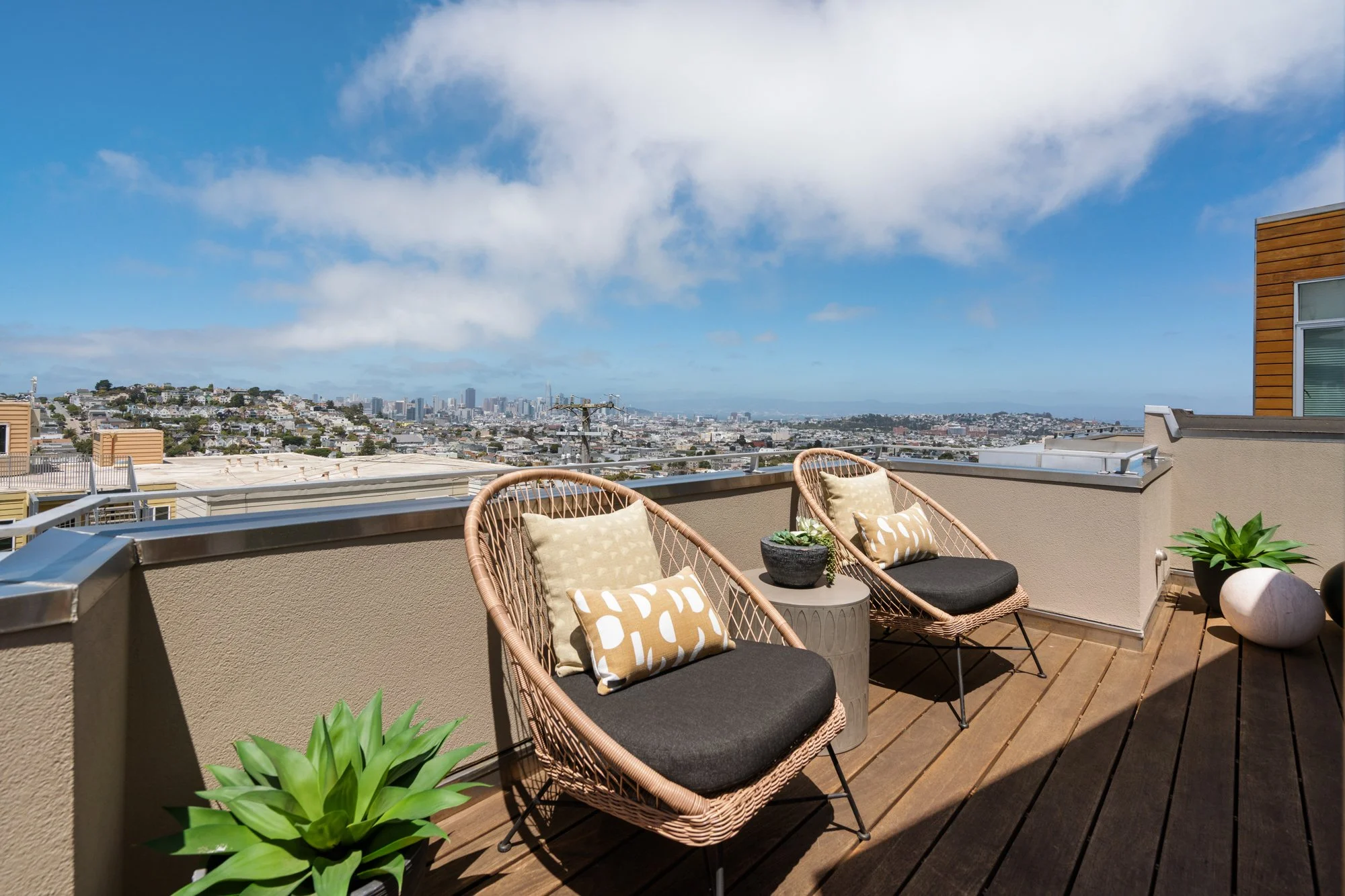 Balcony with two wicker chairs, cushions, and potted plants, overlooking a cityscape under partly cloudy blue sky.