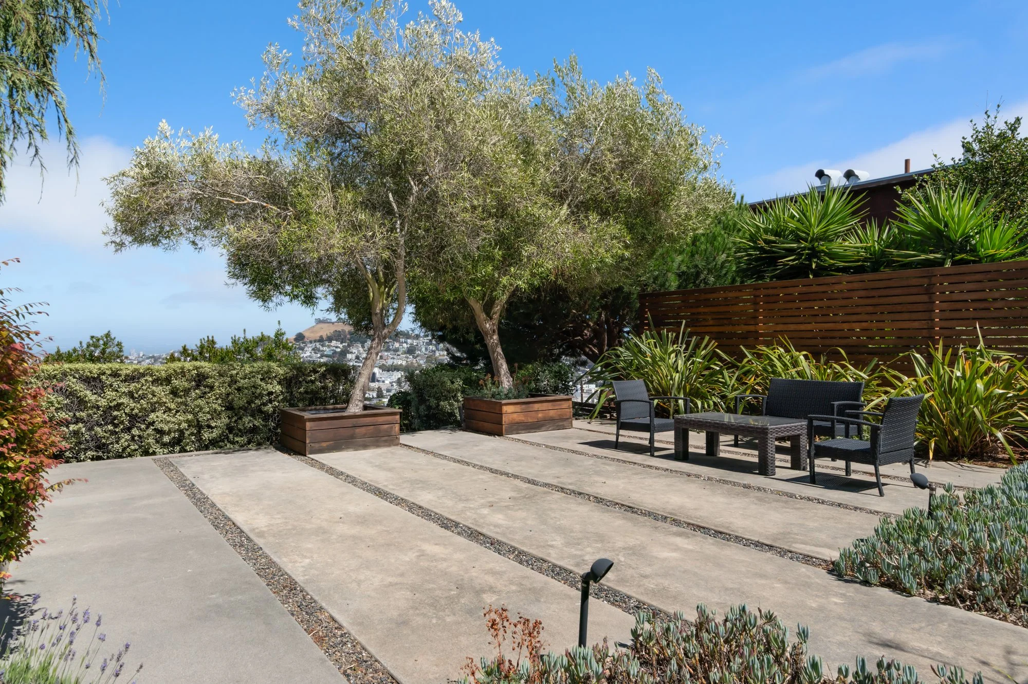 A rooftop patio with a paved area, large potted trees, a modern wooden fence, outdoor seating with black chairs, and lush green plants against a backdrop of a cityscape and hillside under a blue sky.