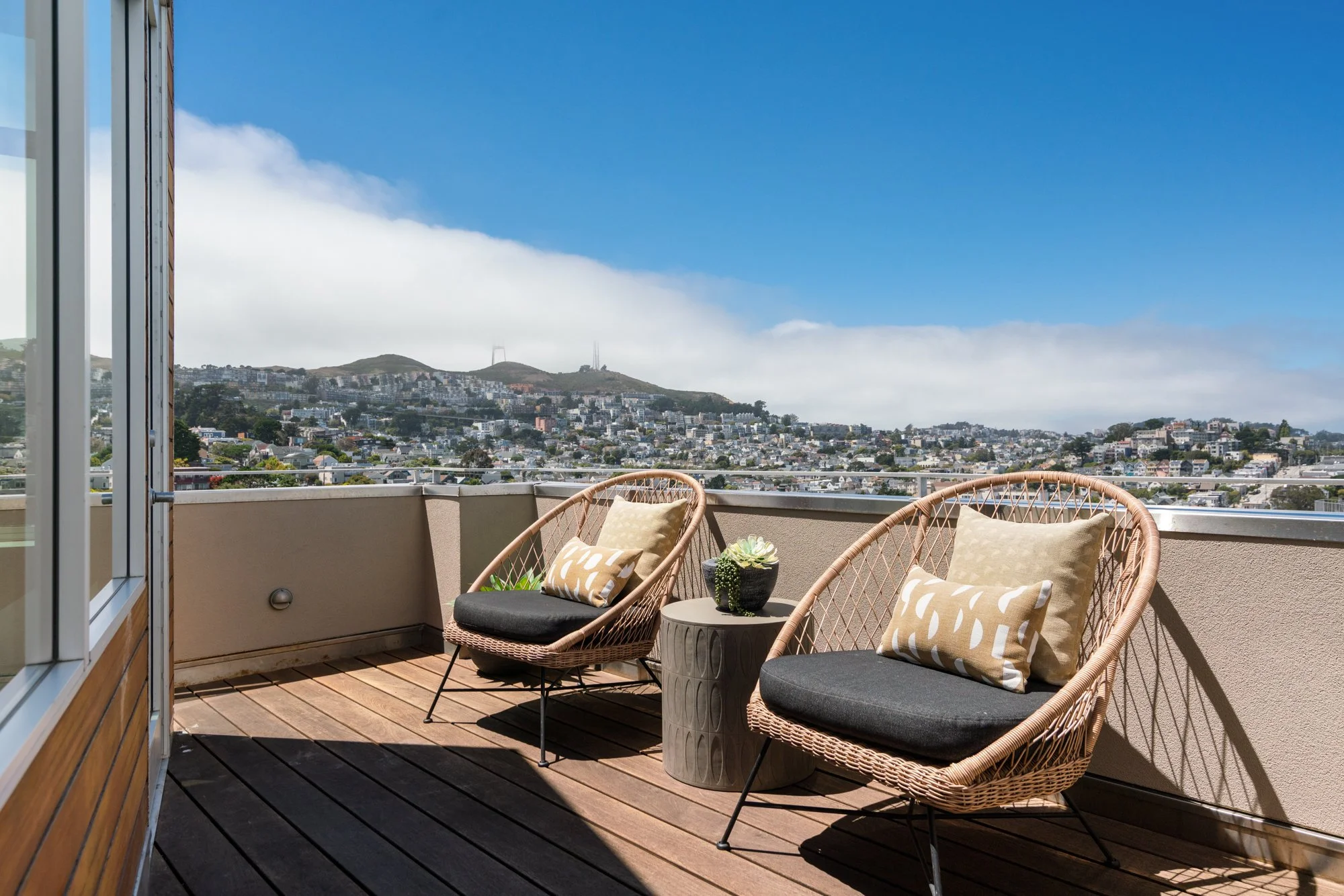 A rooftop balcony with outdoor seating, including wicker chairs with cushions and pillows, overlooking a cityscape with hills in the background and a clear blue sky.