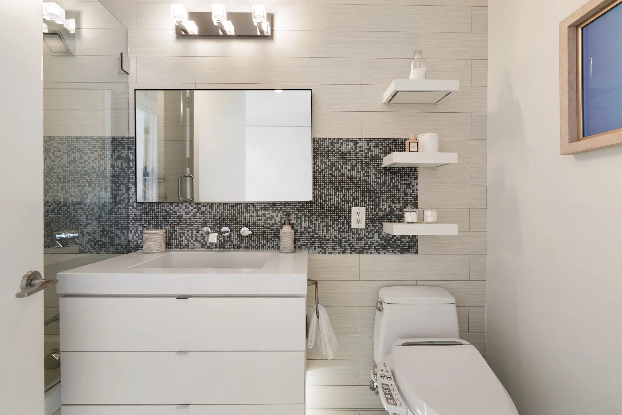 Modern bathroom with beige horizontal tile walls and black and white mosaic tile accent wall behind the sink. Features a large wall mirror, white vanity with laundry basket, toilet with bidet feature, and floating shelves with decorative items and to