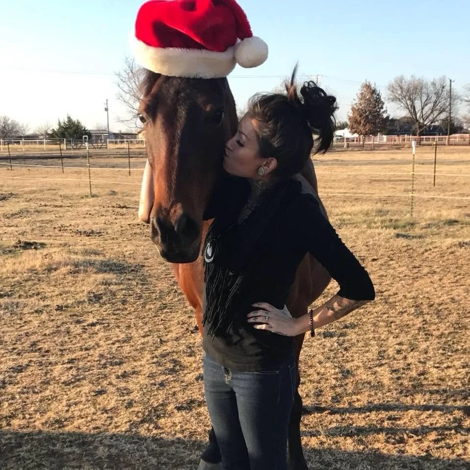 A woman in black top and jeans kisses a horse wearing a Santa hat on its head in an outdoor field.