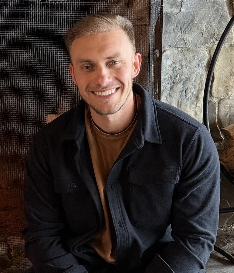 A young man with short, neat hair and a beard, smiling at the camera, wearing a black jacket and beige shirt, sitting indoors with a stone wall and a metal fireplace in the background.