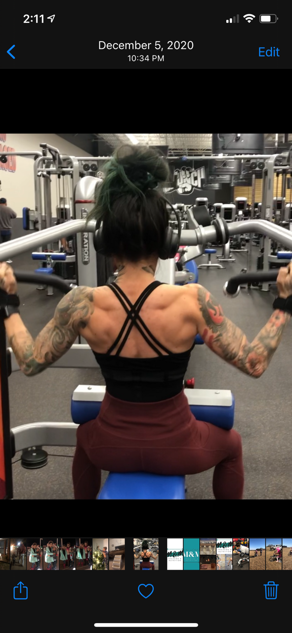 A woman with tattoos on her arms and back, sitting on a blue exercise bench, lifting a barbell at the gym.