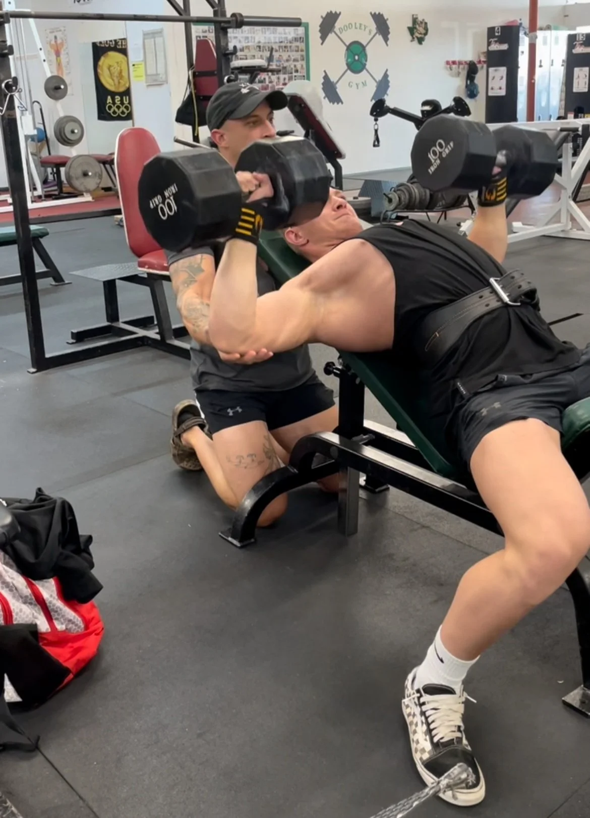 A man is performing a bench press exercise with dumbbells while kneeling on a bench at a gym, assisted by a spotter.