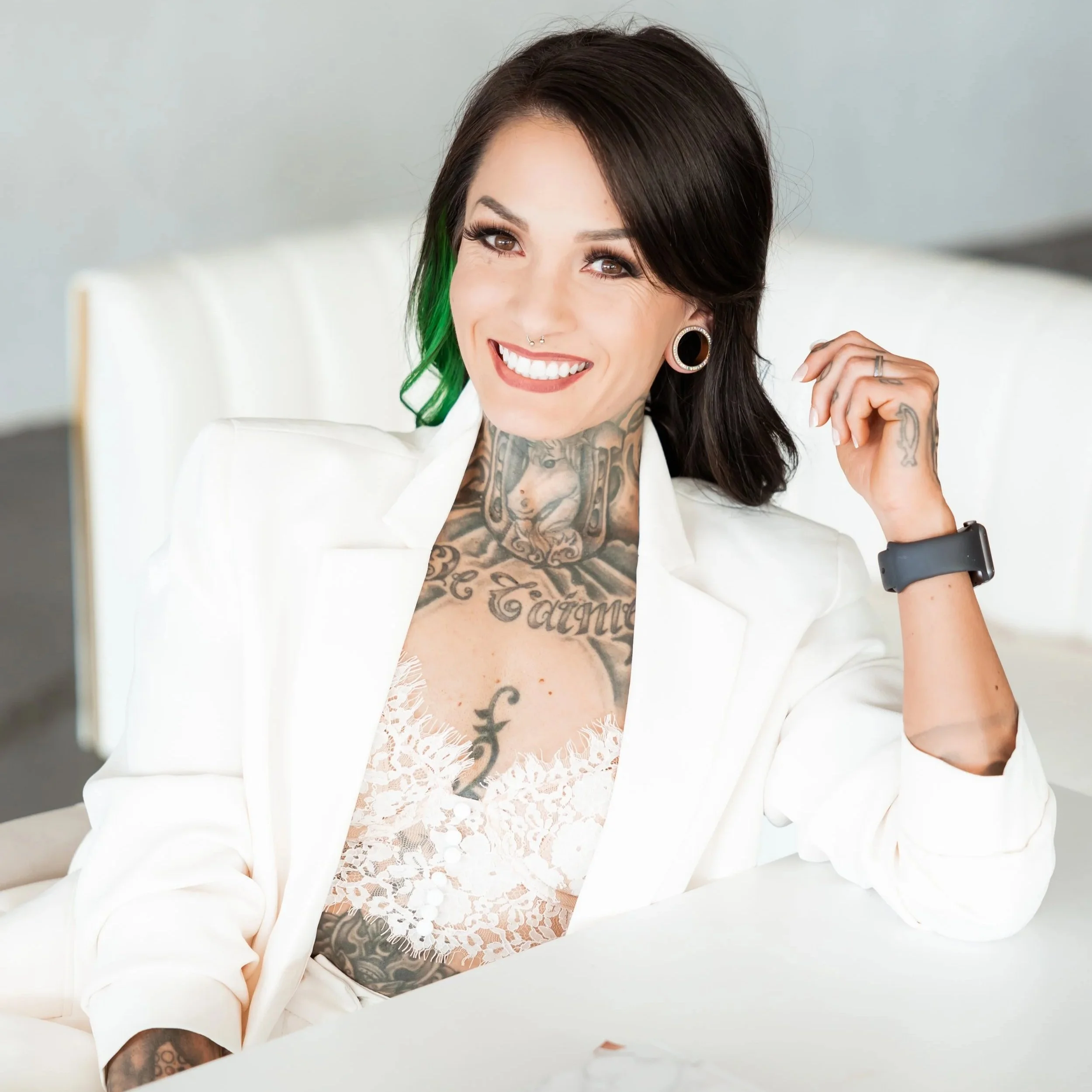 A woman with dark hair and tattoos, smiling, wearing a white blazer and lace top, sitting at a desk with a tablet in front of her.