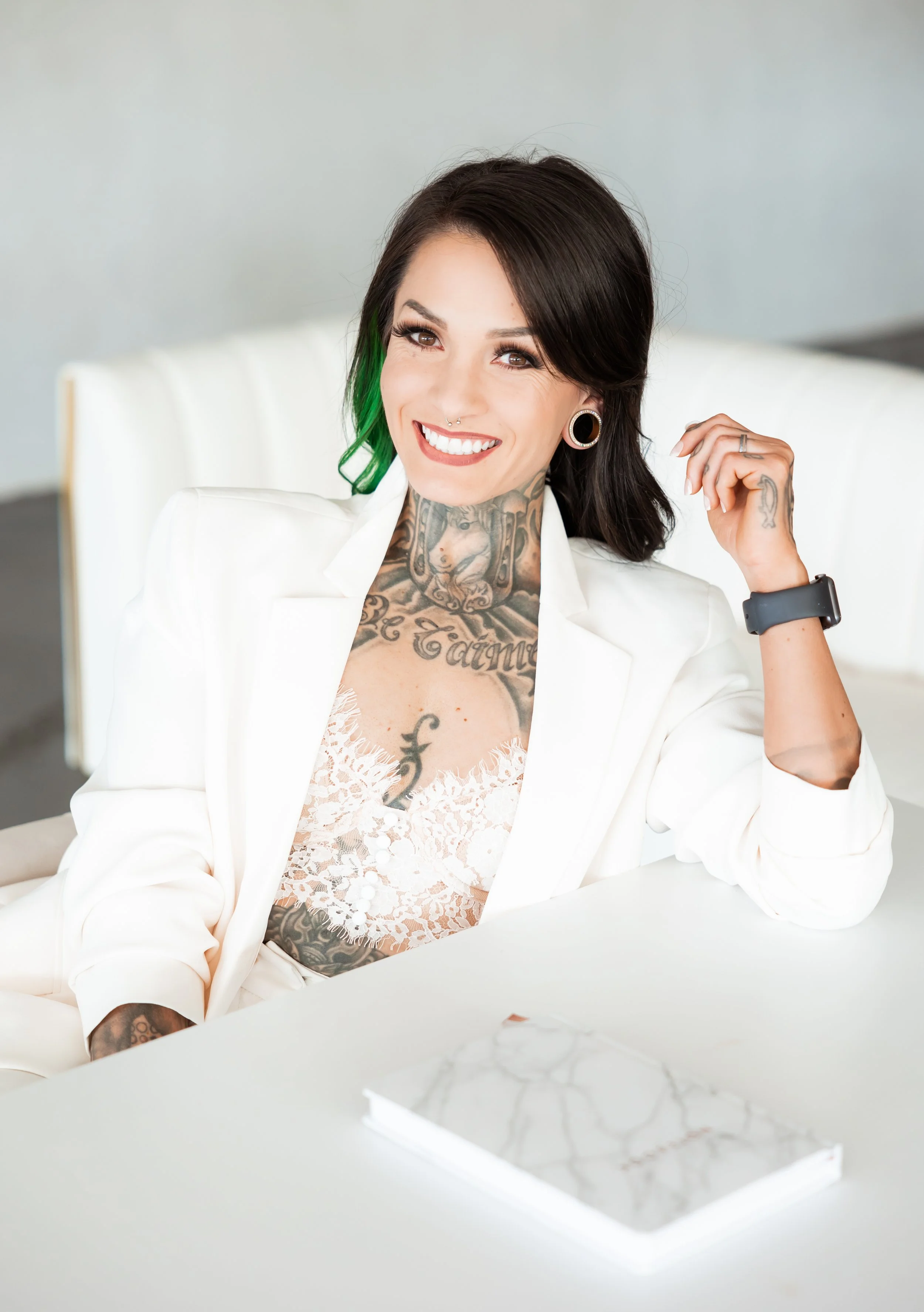 A young woman with dark hair and green tips, smiling, wearing a white blazer, sitting at a white marble table with a marble-patterned tray in the foreground, in a bright room. djvanillaface dj madson