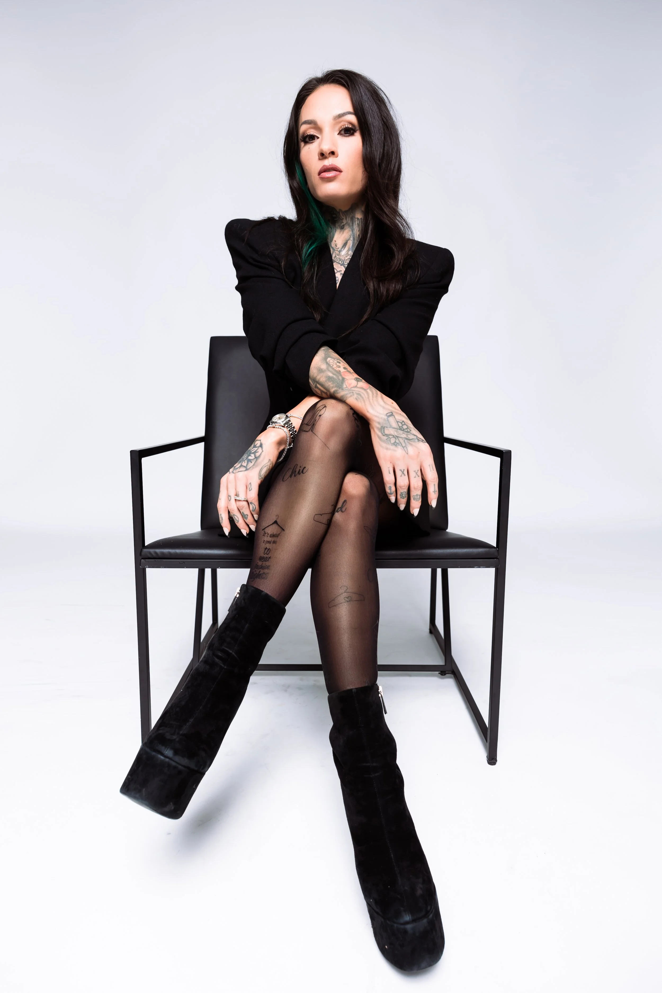 A woman with long dark hair and tattoos, sitting on a black chair, wearing a black blazer, sheer tights with tattoos, and black high boots, in a studio with a plain light gray background. djvanillaface dj madson