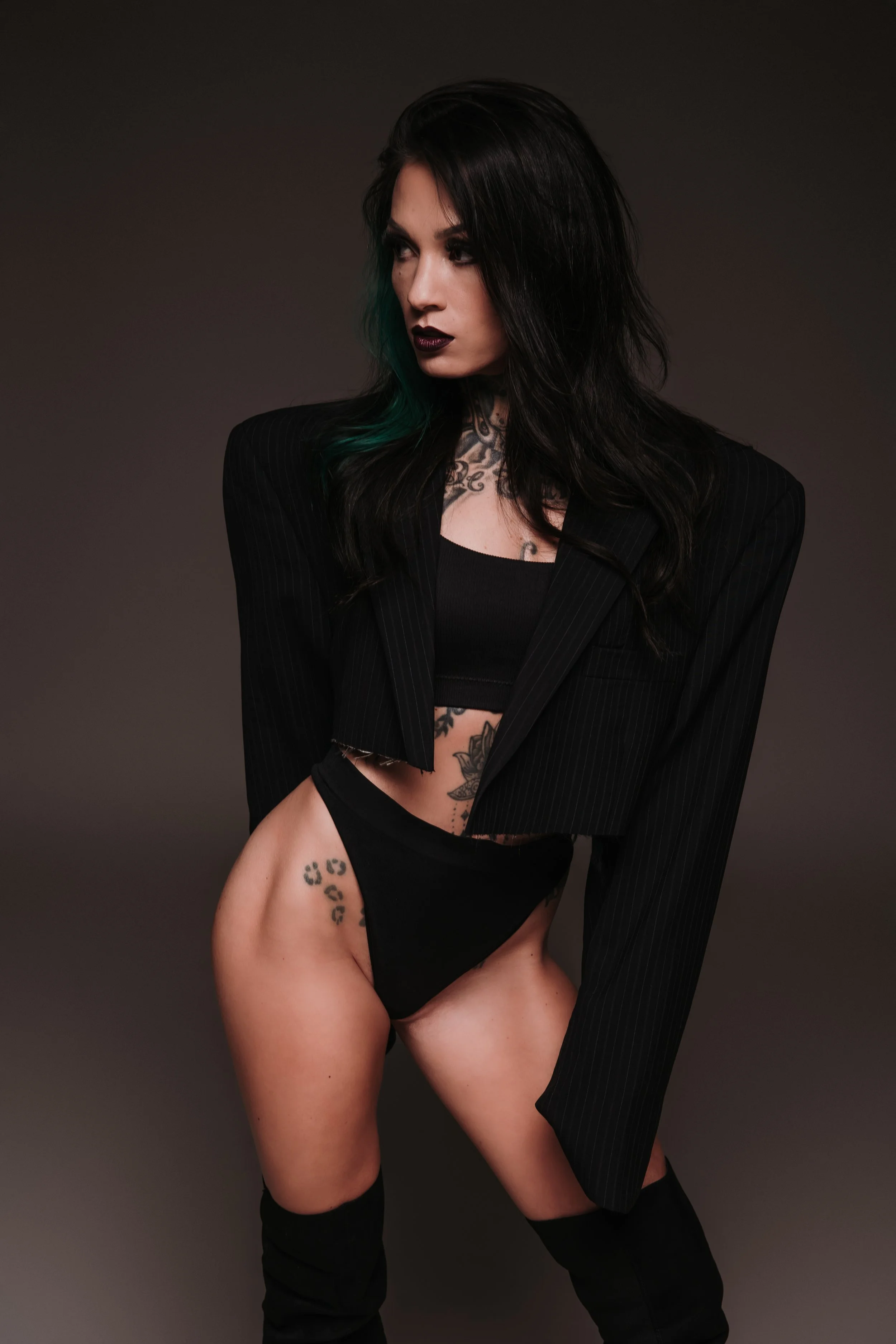 A woman with black hair and tattoos posing against a dark background, wearing a black pinstripe blazer, black tank top, black underwear, and thigh-high black boots, with a confident expression. djvanillaface dj madson