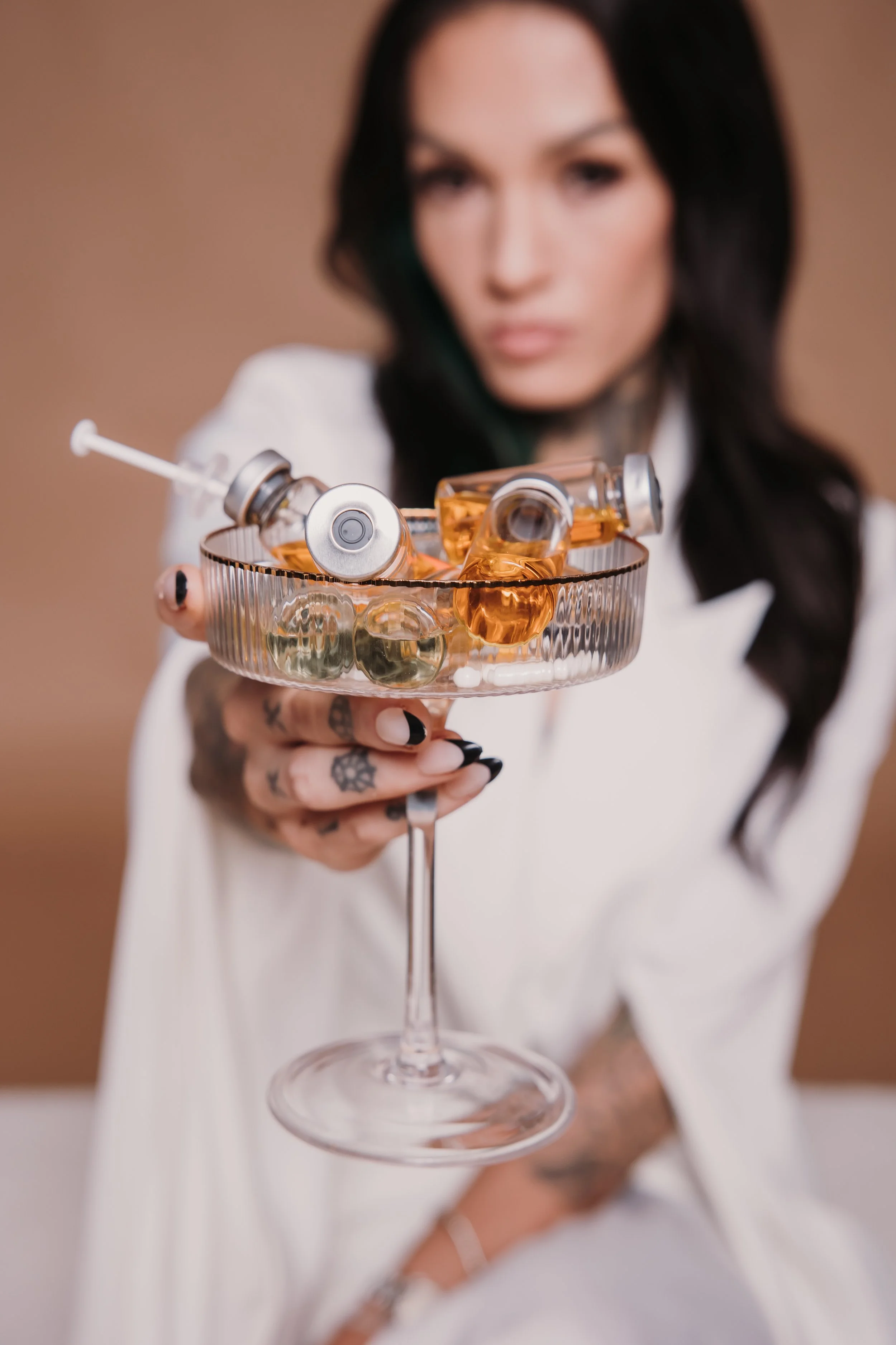 A woman with black hair and tattoos holding a glass of liquor with ampoules and a syringe inside, blurred background.djvanillaface dj madson