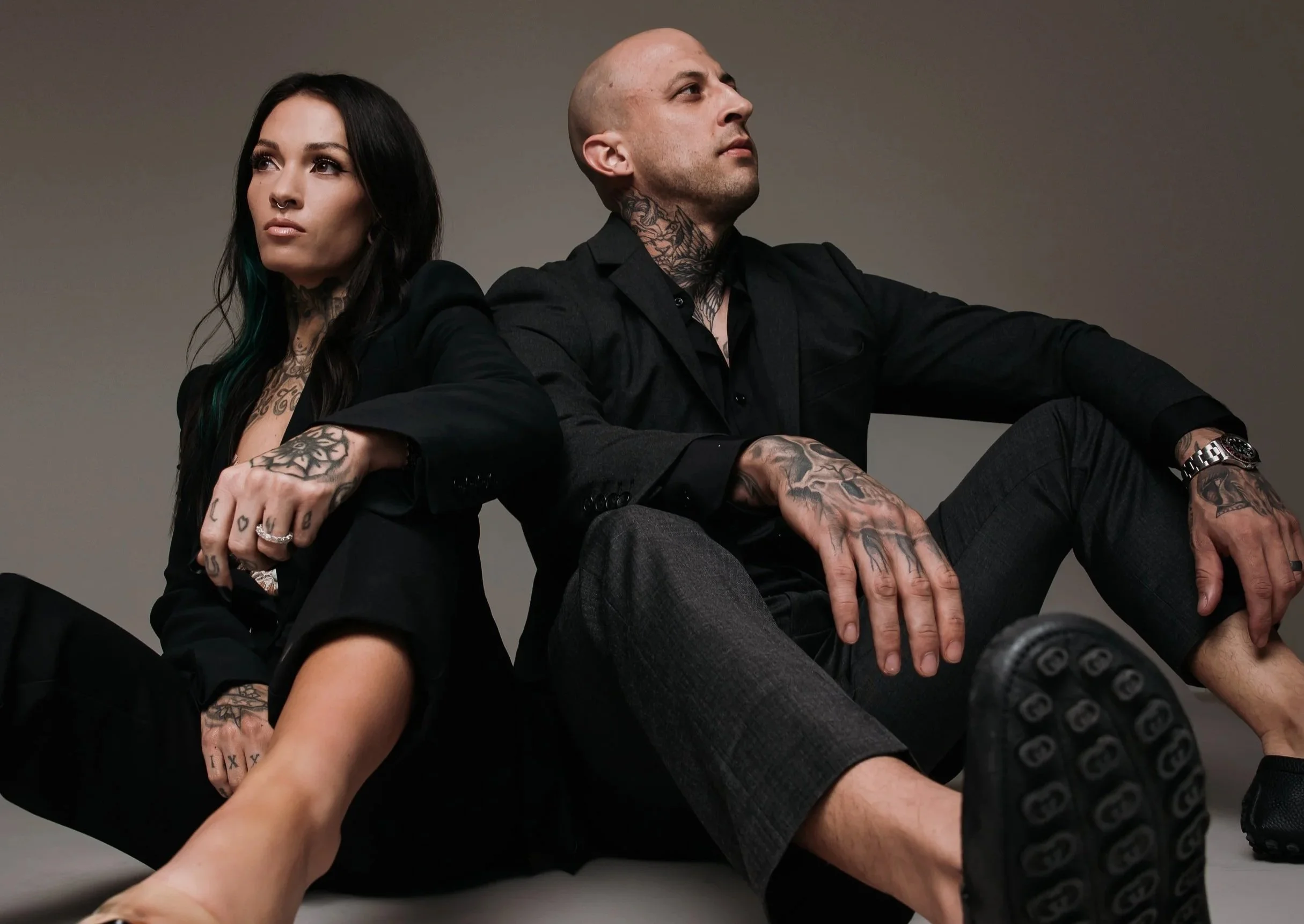 A fashion photo of a man and a woman sitting back to back. The woman has dark hair with green tips, tattoos, a septum piercing, and a confident expression. The man has a shaved head, tattoos on his neck and hands, and is looking upward with a serious expression. They are dressed in black.