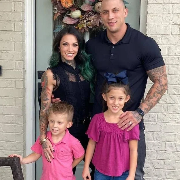 A family of four standing in front of a door decorated with a flower wreath, smiling at the camera. The woman has dark hair with green tips, and the man has tattoos on his arms. Two children, a girl in a pink shirt with a large blue bow in her hair and a boy in a pink polo, stand in front of them.