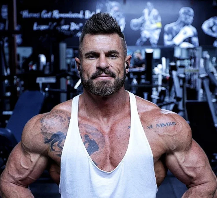 A muscular man with a beard and styled hair in a white tank top, standing in a gym with workout equipment and muscle artwork in the background.
