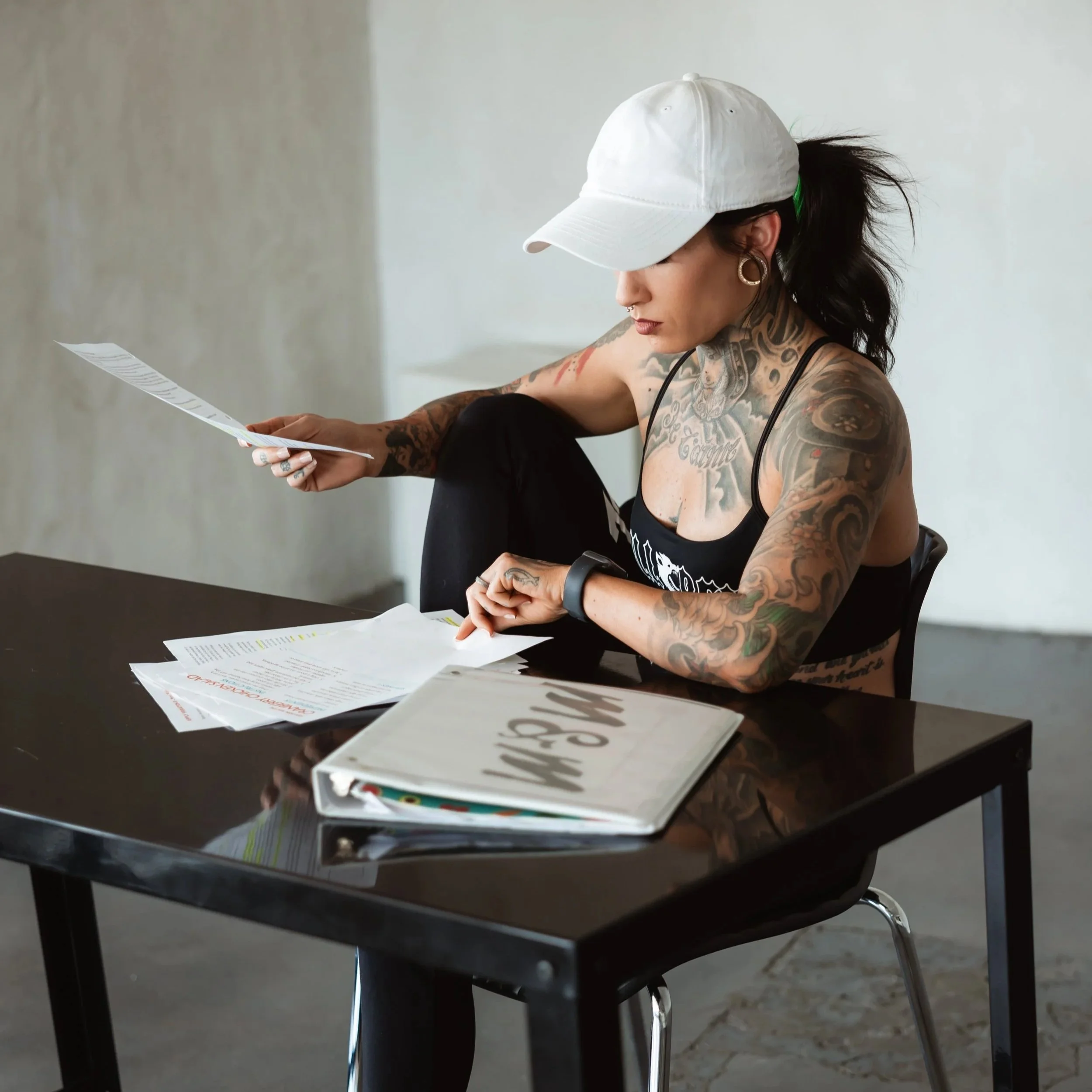 A woman with numerous tattoos wearing a white cap and earrings, sitting at a black table reviewing papers, with additional papers on the table. djvanillaface dj madson