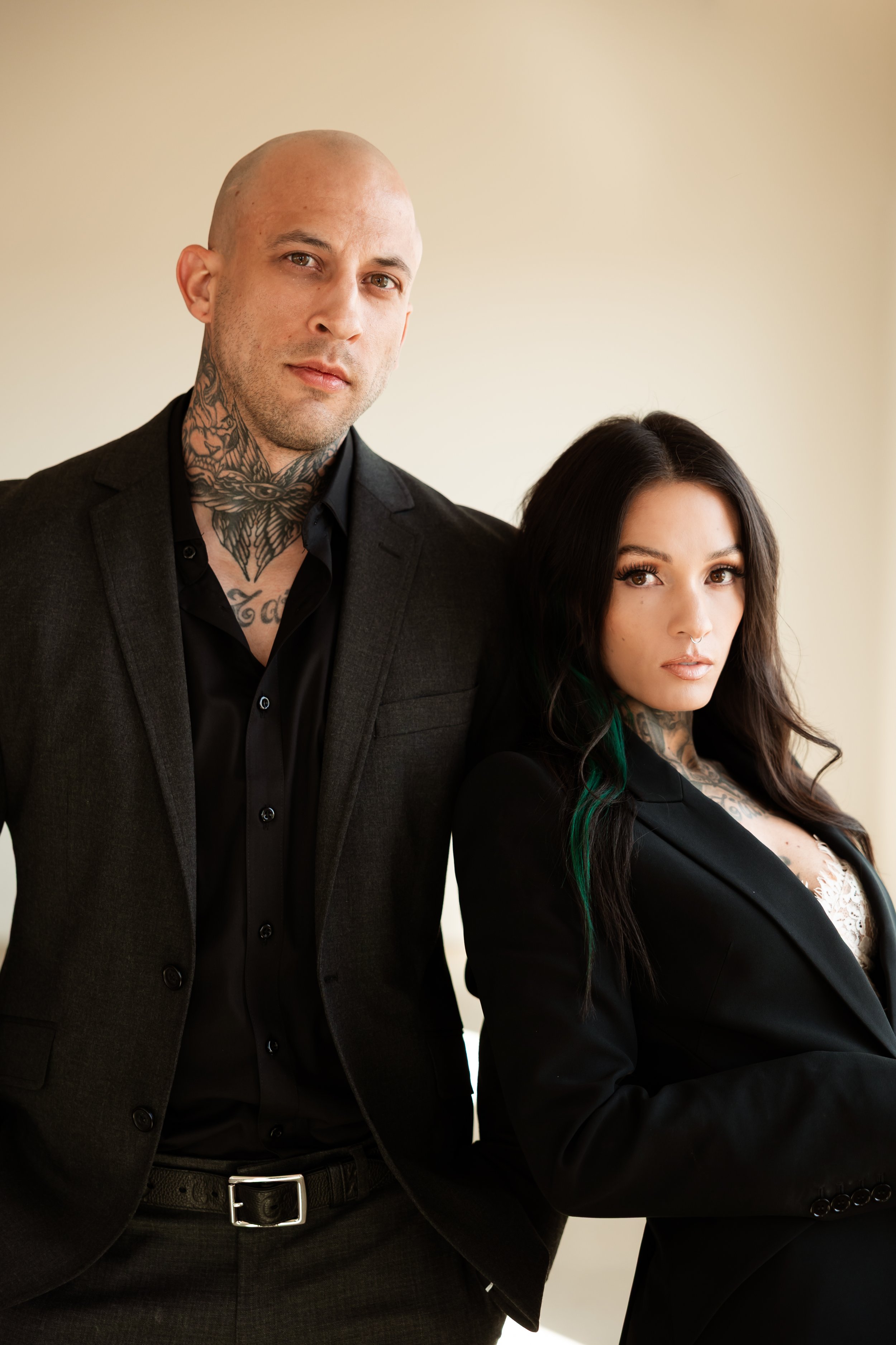 A man and woman dressed in black suits posing together against a plain, light-colored background.