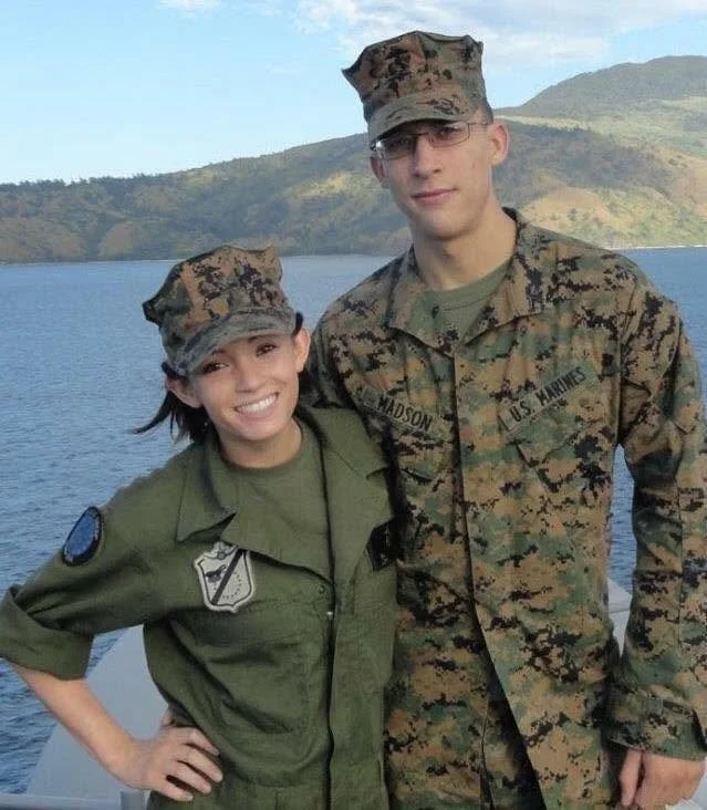 Two U.S. Marines in uniform standing outdoors near a body of water with hills in the background, smiling at the camera.