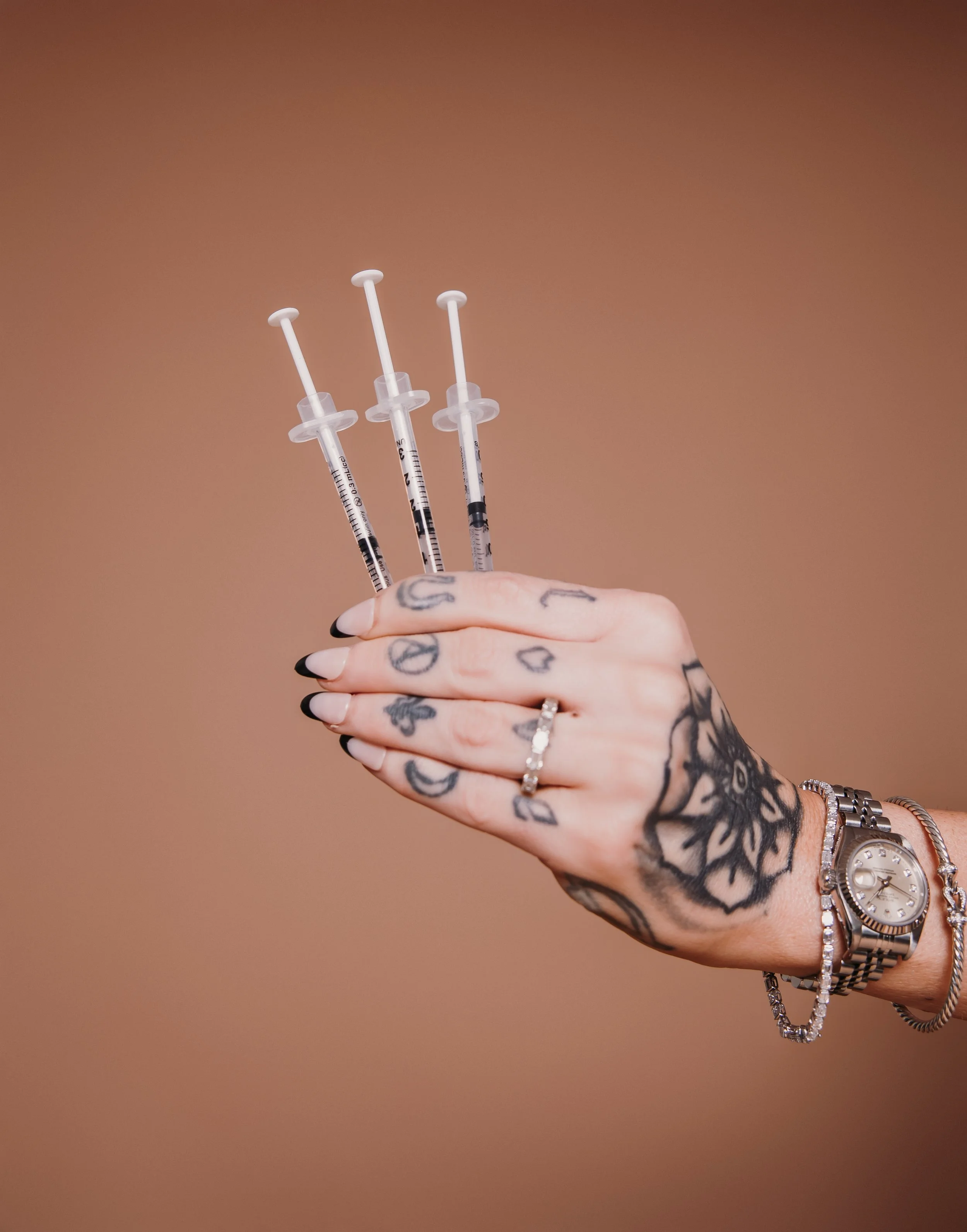 Close-up of a person's hand holding three syringes with needles pointed up, tattoos visible on the hand and wrist, a ring on the finger, and a silver watch and bracelet on the wrist.